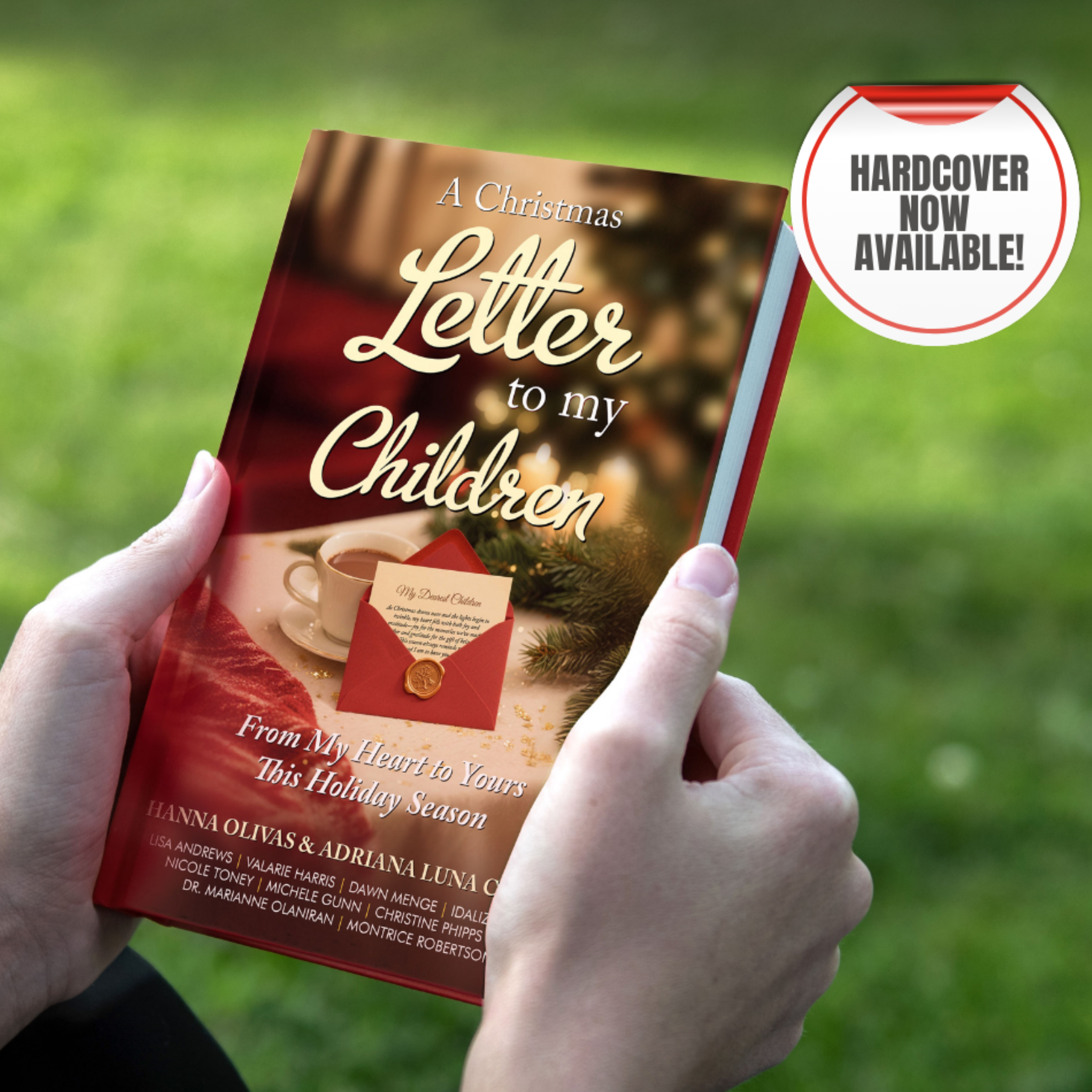 A person holding a Christmas-themed book titled "A Christmas Letter to my Children" with a blurred background of green grass. The cover features a cup of hot chocolate, a red envelope with a letter, and a Christmas tree. There is a white sticker on the top right corner that says "Hardcover now available!"