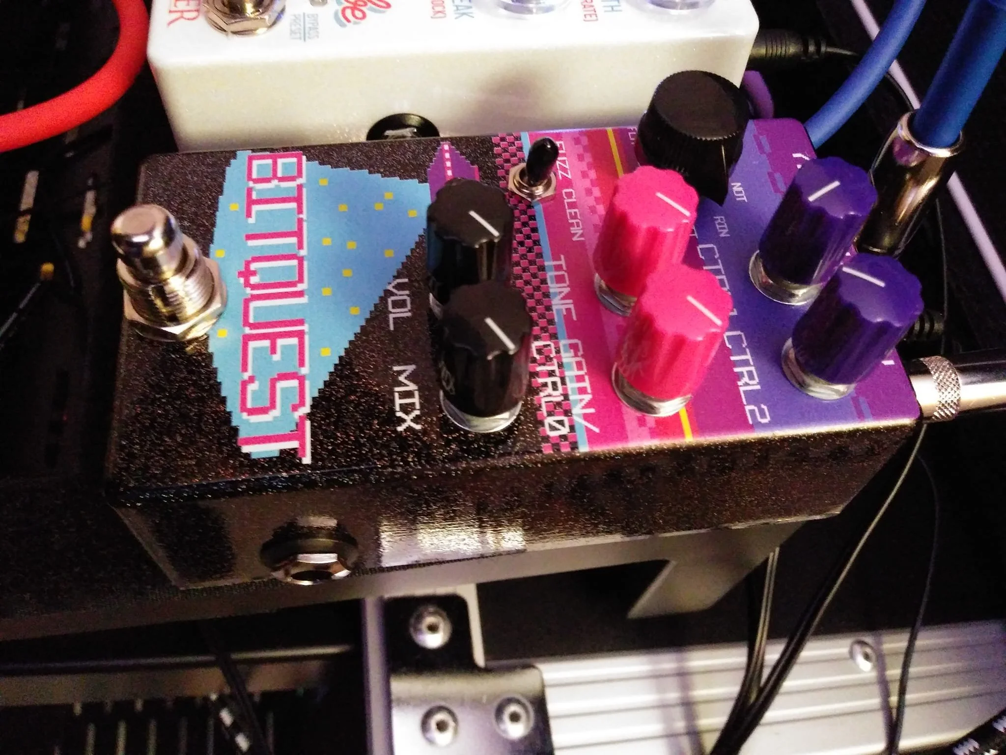 Close-up photo of a section of a colorful audio effects pedal with various control knobs and switches, placed on a guitar amplifier with cables connected.