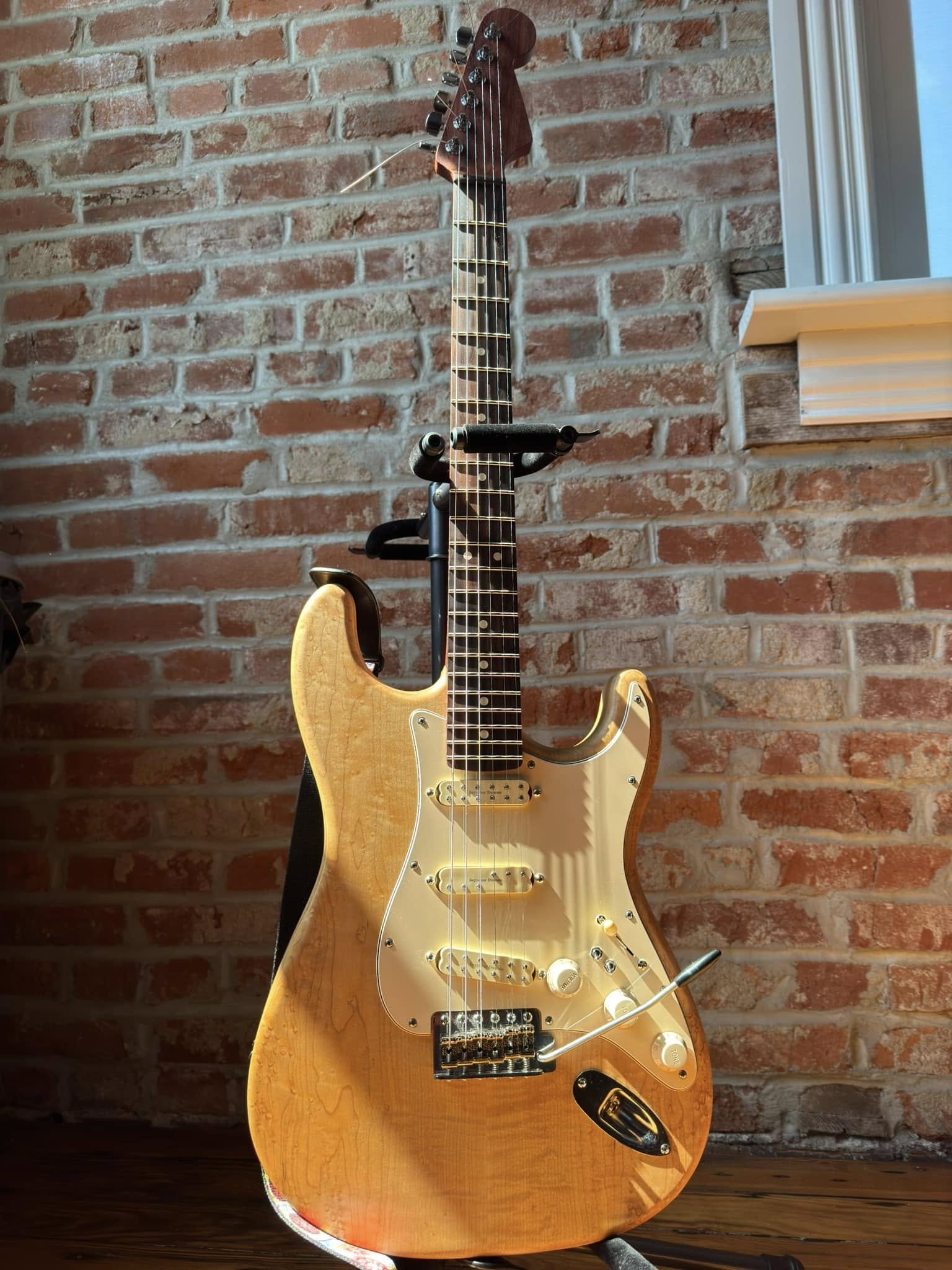 A cream-colored electric guitar with a wooden finish, placed on a guitar stand against a brick wall.