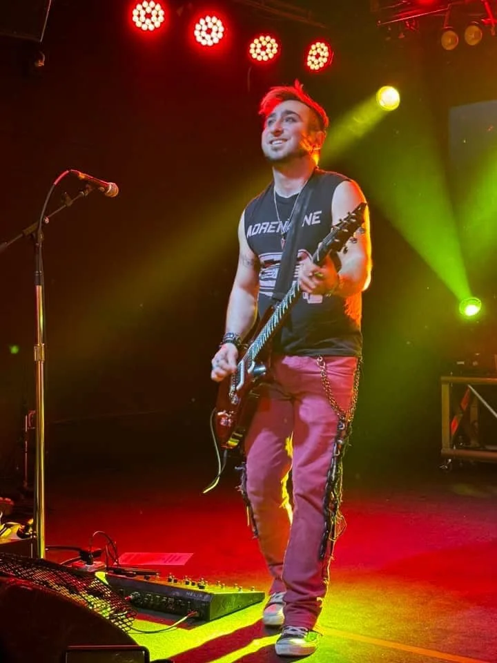 A young male musician playing an electric guitar on stage, smiling, with colorful stage lights overhead, microphone stand to his left, and music equipment on the floor.