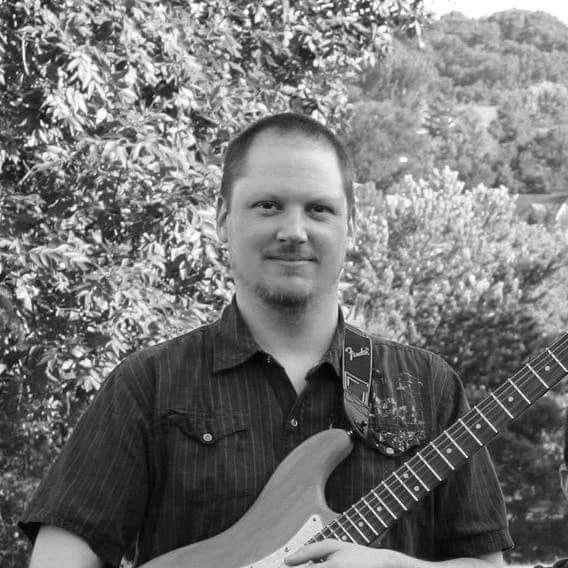 A man holding an electric guitar outdoors, with trees and a hillside in the background, in black and white.