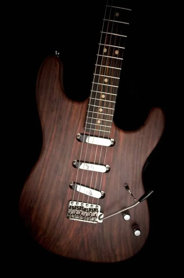A close-up of a wooden electric guitar with a dark finish, white pickups, and a tremolo bridge.