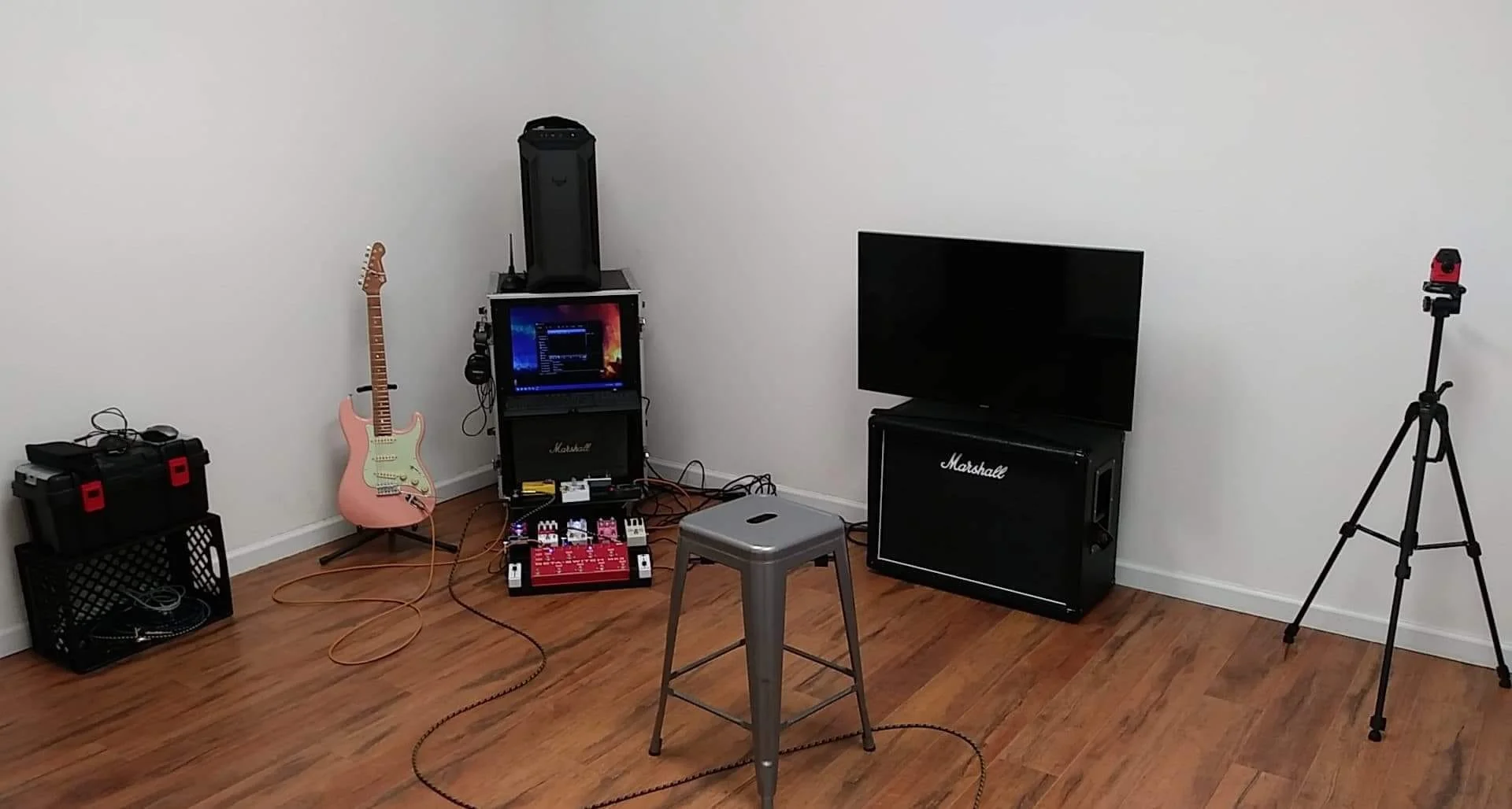 Music practice or recording setup in a room with a white wall and hardwood floor, including a pink electric guitar, a computer on a stand, a Marshall amplifier, a large TV screen, small stool, and various audio equipment and accessories.