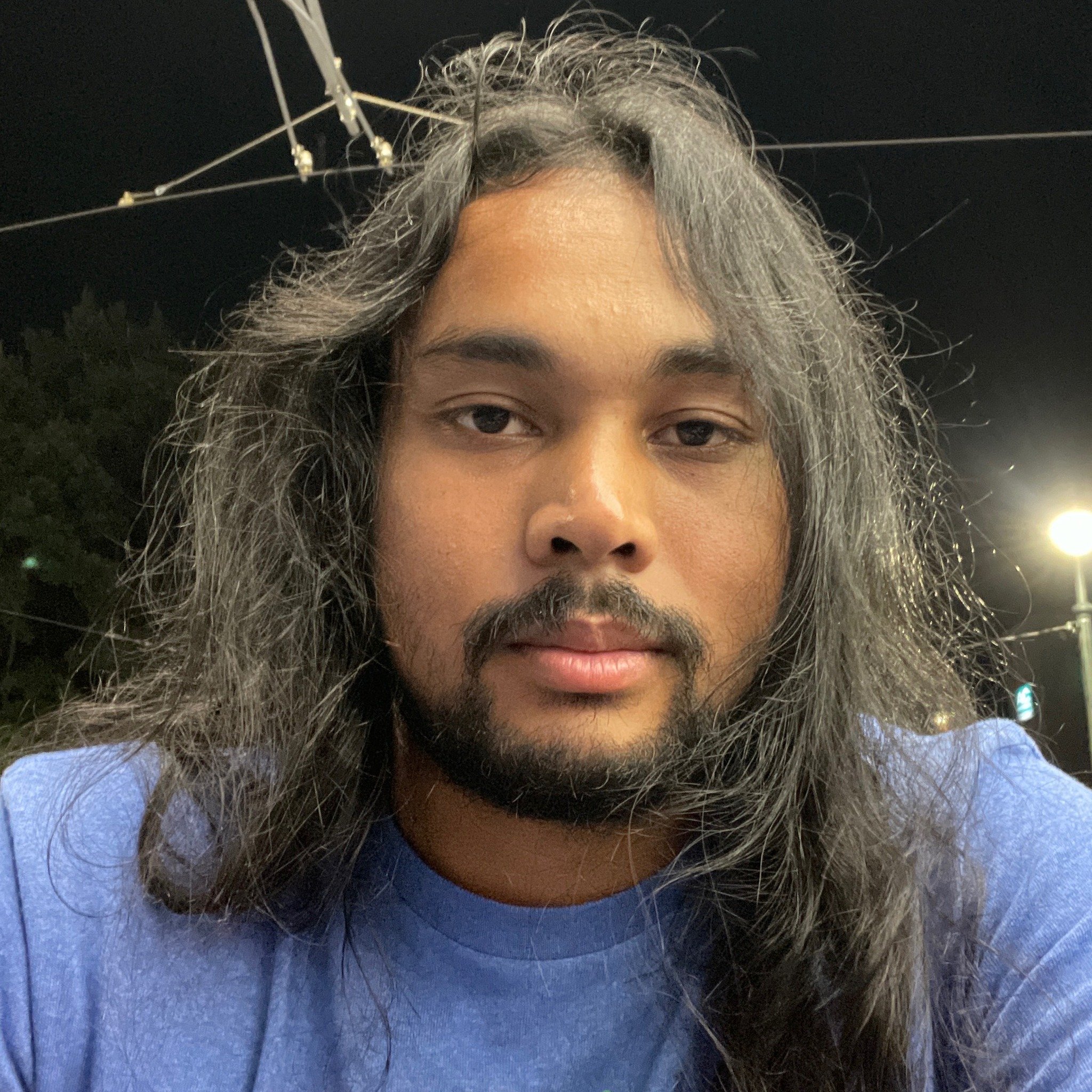 Close-up of a person's face with long gray hair, wearing a dark blue top, with a black background and some metal structures above the head.