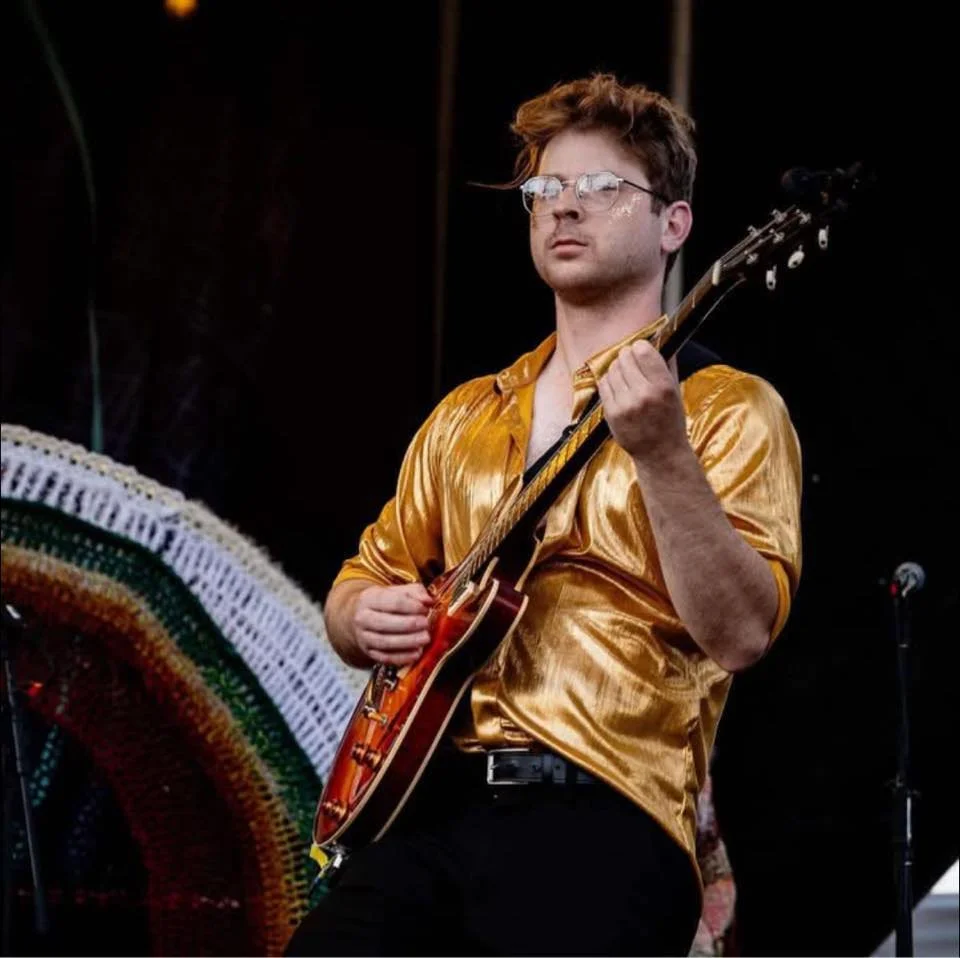 A young man with glasses and messy hair playing an electric guitar on stage, wearing a shiny gold shirt.