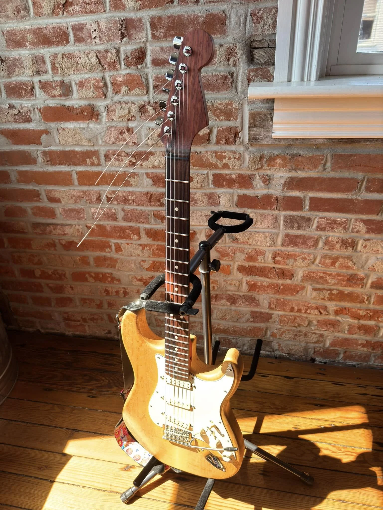 Sunlit electric guitar with a light wood finish on a stand, against a brick wall and wooden floor, near a window.