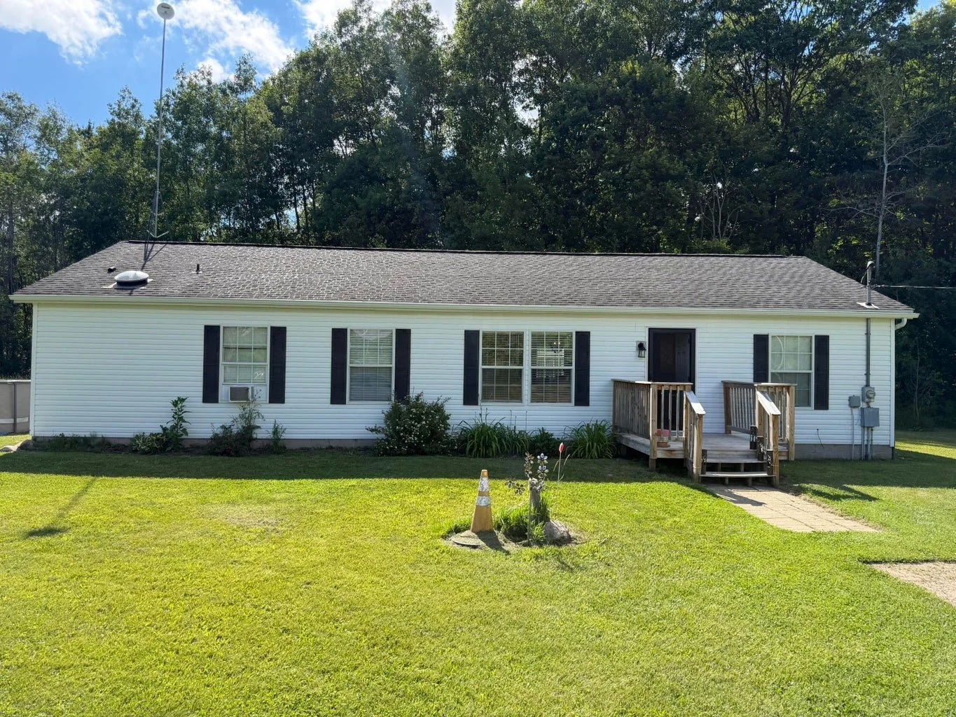 A single-story house with white siding, black shutters, a gable roof, and a small wooden porch with stairs in the front. The house has 5 windows, with one air conditioning unit in a window on the left. The yard is grassy with some plants and a small flower bed in front. There's a concrete pathway leading to the porch, and the background features a line of tall trees under a blue sky.