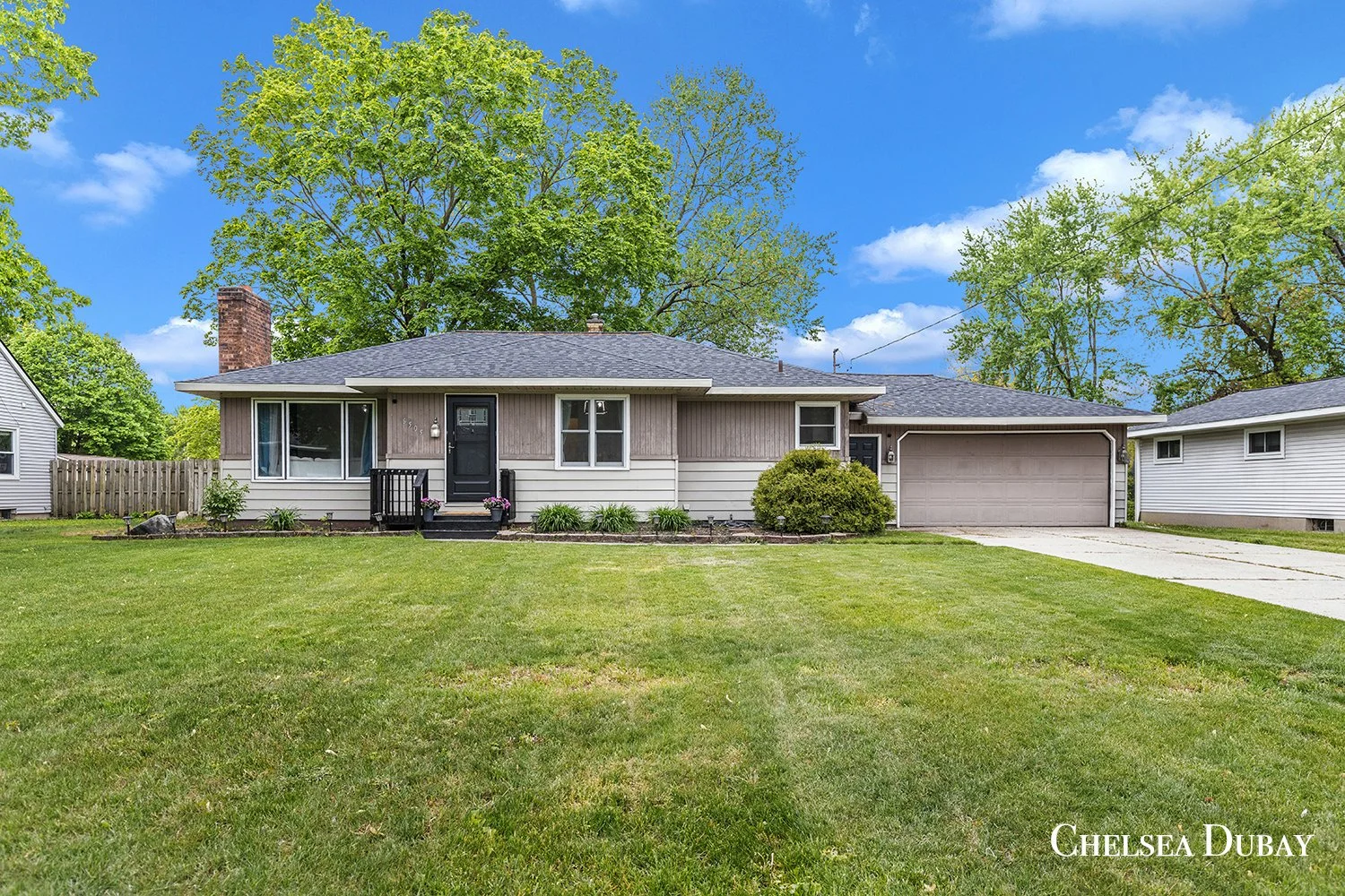 A single-story house with a gray exterior, black front door, and an attached garage, surrounded by a well-maintained lawn and trees with green leaves under a bright blue sky. Jenison, west Michigan real estate, realtor, 5 star review, top agents in w