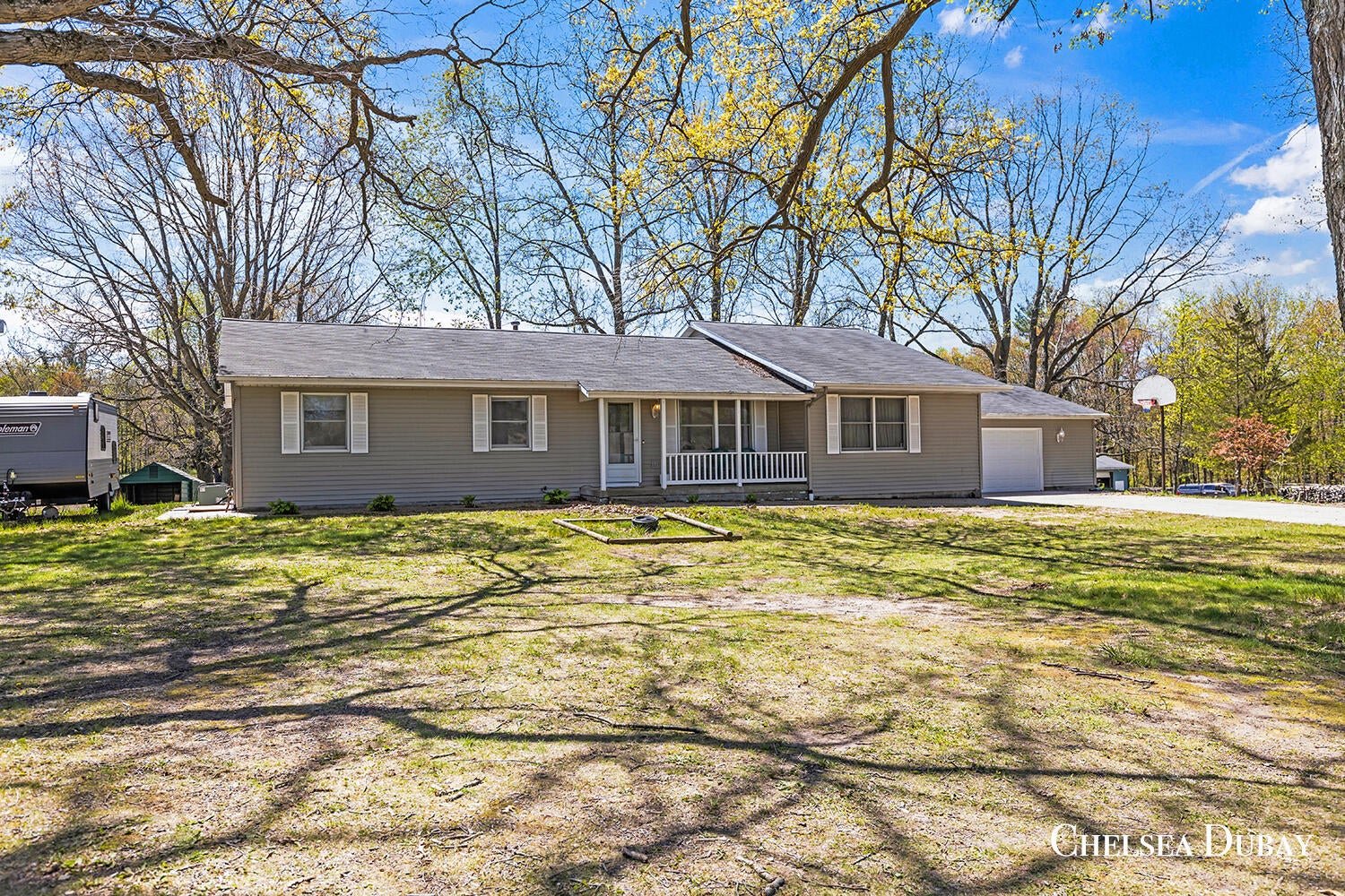 A single-story house with a light-colored exterior, front porch, and attached garage, surrounded by a spacious yard with trees and shadows on the ground. A basketball hoop is visible near the driveway on the right side. rockford, mi