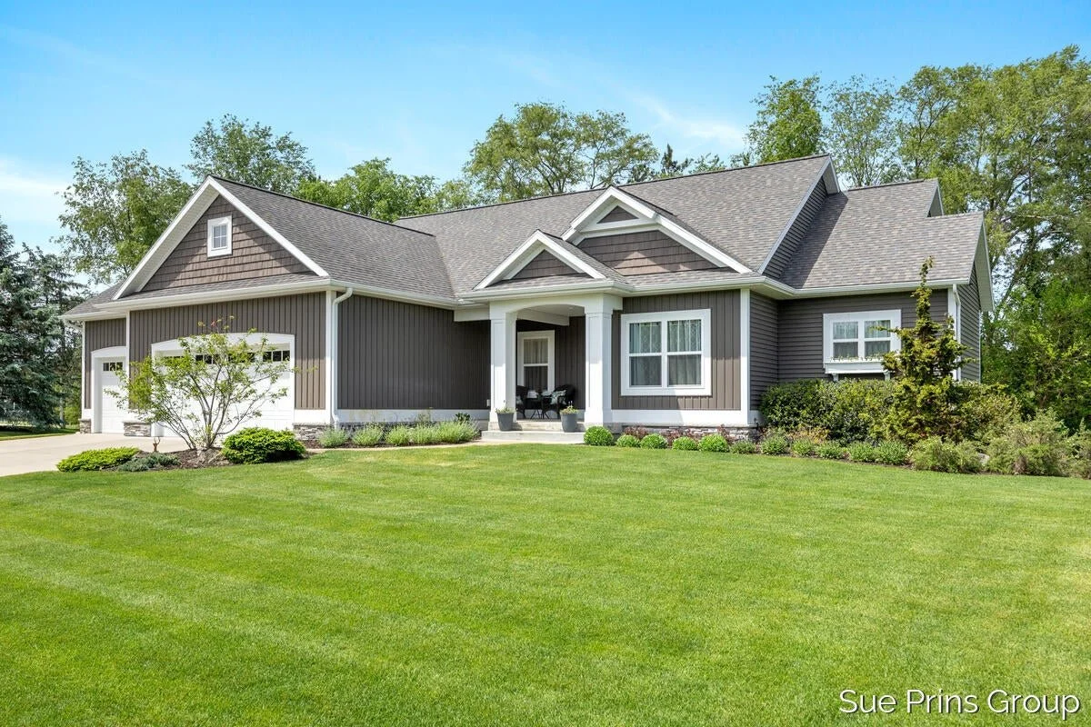 A modern suburban house with gray exterior walls, white trim, and a gabled roof, surrounded by a well-maintained lawn, trees, and shrubs. Hudsonville, west Michigan real estate, realtor, 5 star review, top agents in west Michigan.