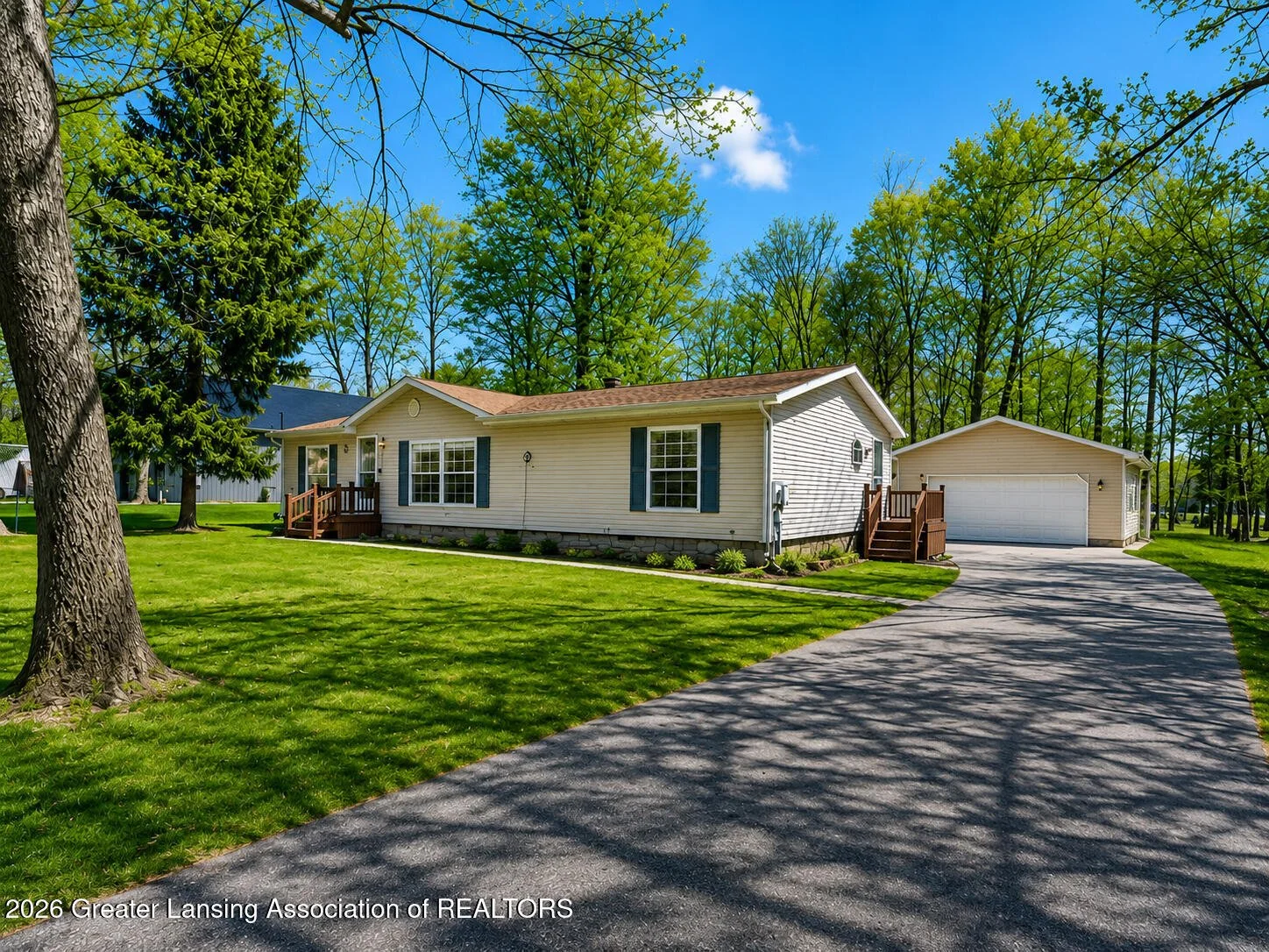 A single-story house with white siding and blue shutters, a small front porch with steps, and a detached garage on a curved asphalt driveway. The house is surrounded by a well-kept lawn and tall trees under a bright blue sky.