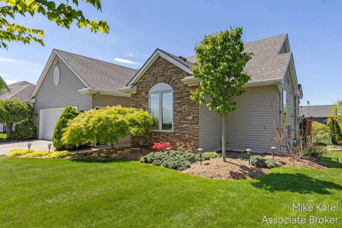 Front yard of a house with a well-maintained lawn, various trees, shrubs, and a stone and siding exterior, under a clear blue sky.