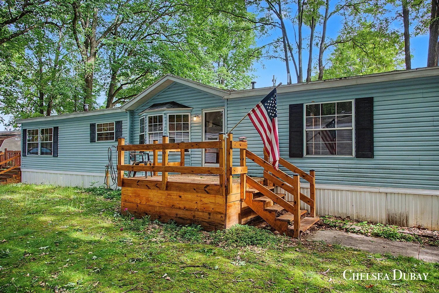 Backyard of a blue manufactured home with a wooden deck, American flag, and trees in the background. for sale Sparta, mi. West Michigan real estate