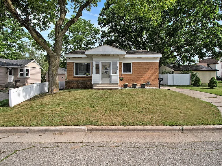A single-story brick house with a white front porch and steps, surrounded by a well-maintained lawn and large trees in a suburban neighborhood.
