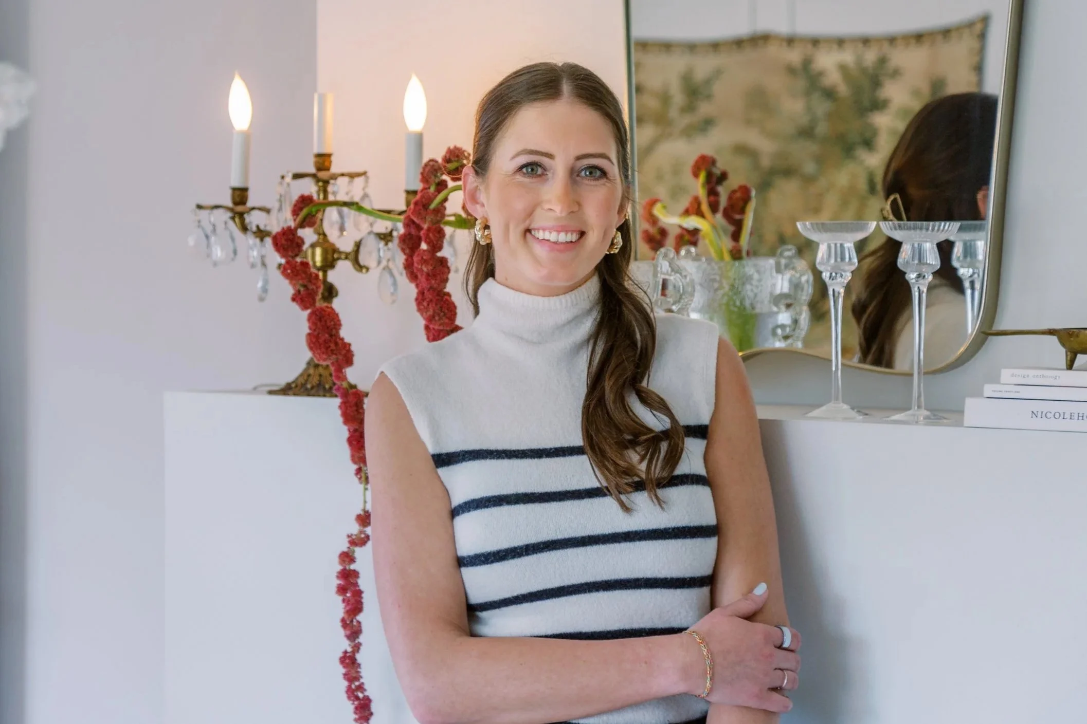 A woman with long brown hair in a loose braid, wearing a sleeveless white turtleneck sweater with black stripes, smiling while standing in a room with decorative items and a mirror.