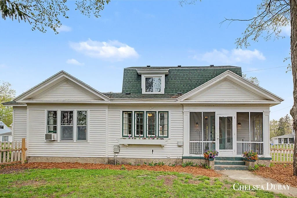 A white, two-story house with a screened porch and a small front yard with green grass and flower pots.