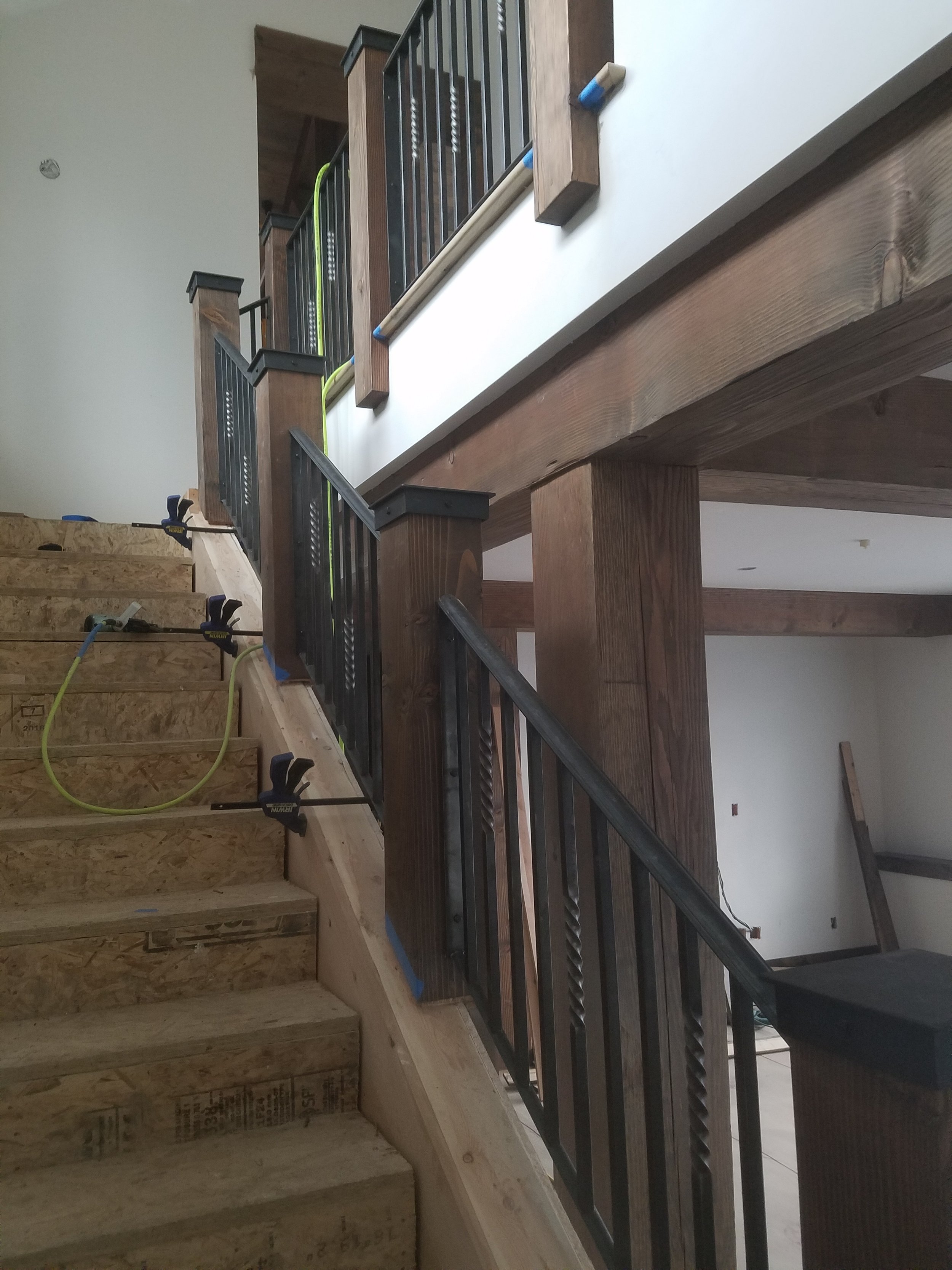 Interior of a house under construction showing wooden stairs, a black metal railing, and a second-story balcony with wooden posts and black and green accents.