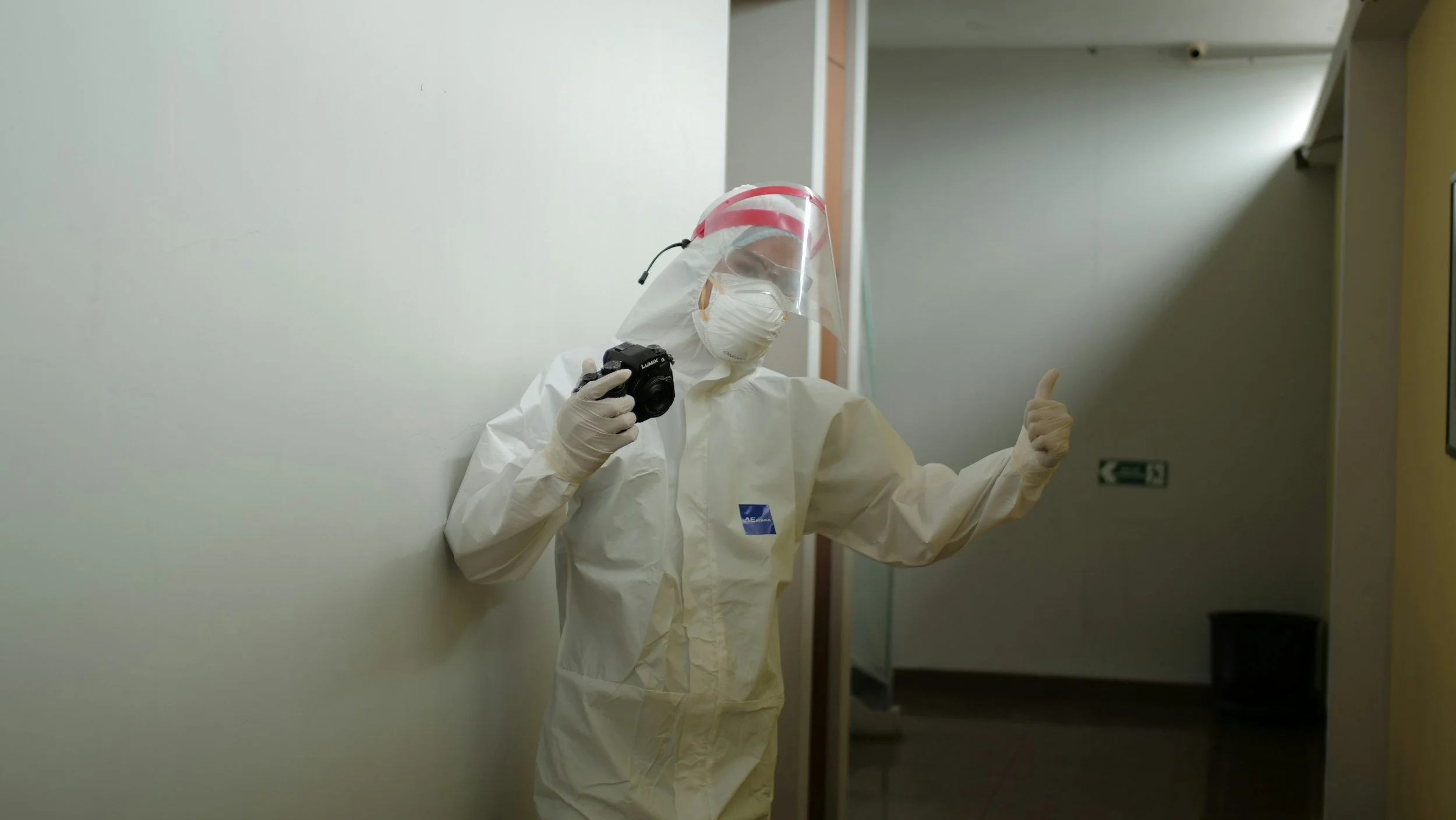 Person wearing full protective gear including a face shield, mask, gloves, and a white suit, holding a camera and giving a thumbs up while standing in a hallway.