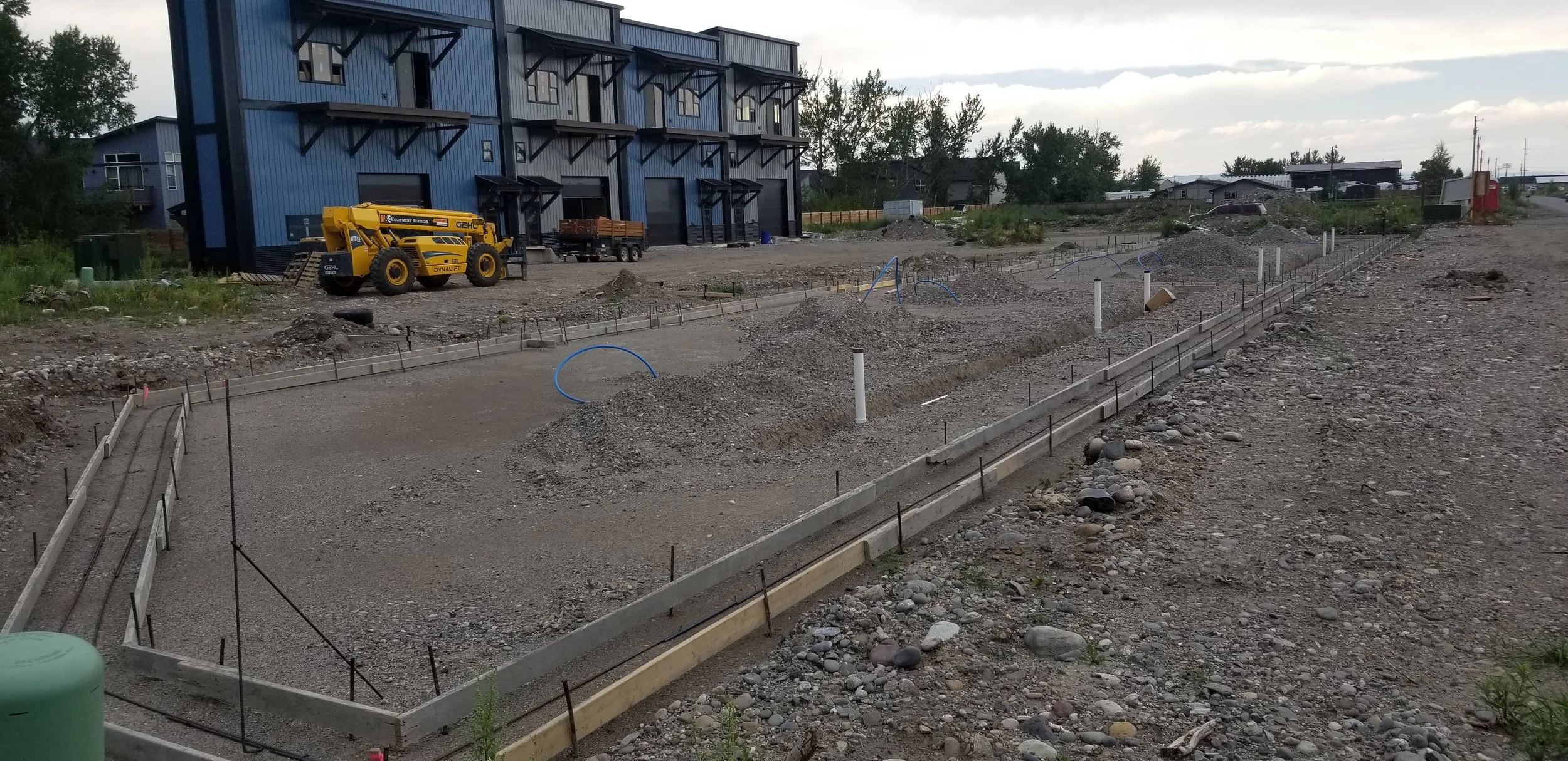 Construction site with foundation and piping in progress, blue building in background, and construction equipment on the site.