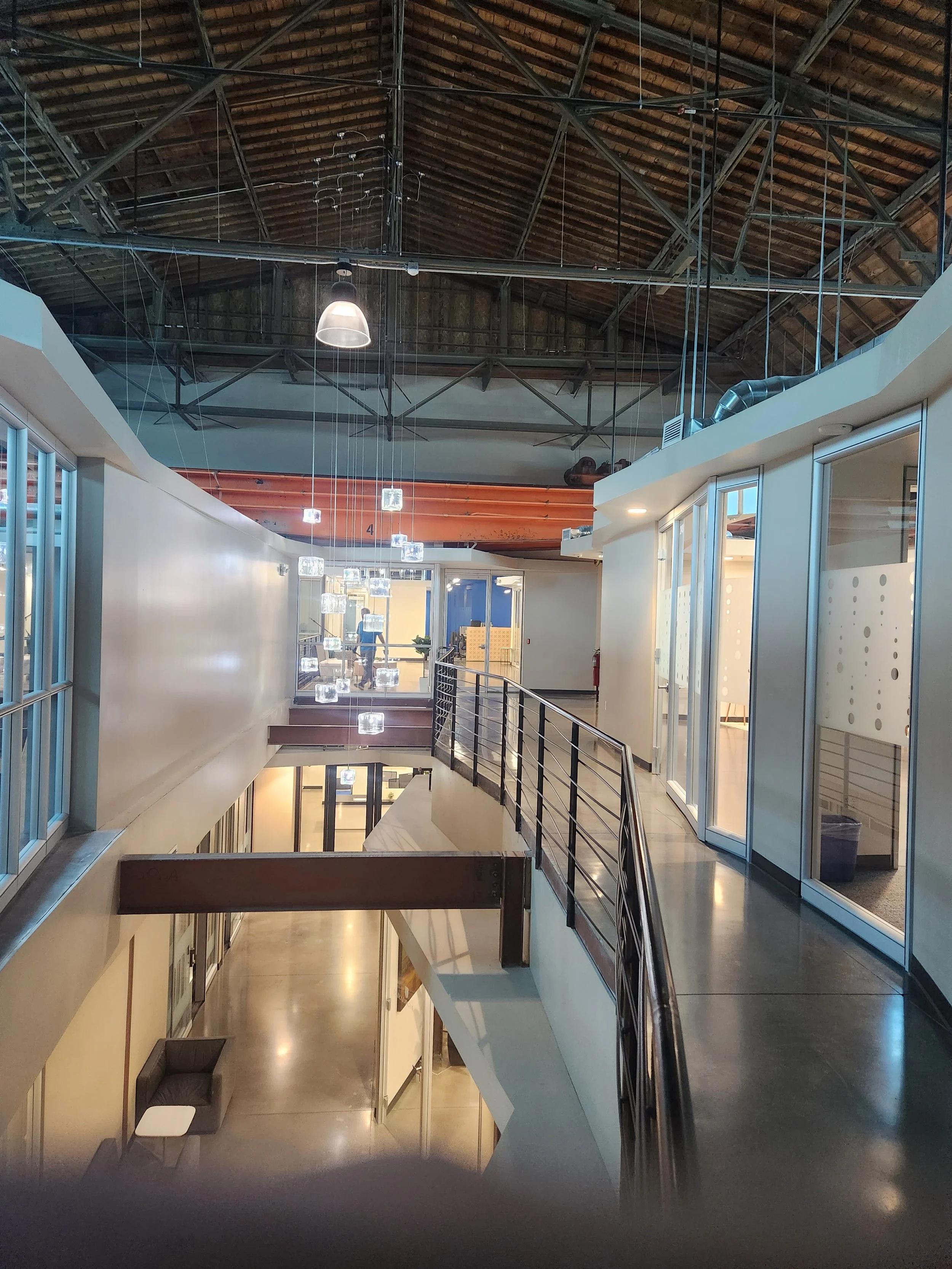 Interior view of a modern office with open corridors, glass offices, a staircase, and decorative hanging light fixtures, with a high wooden ceiling and visible structural beams.