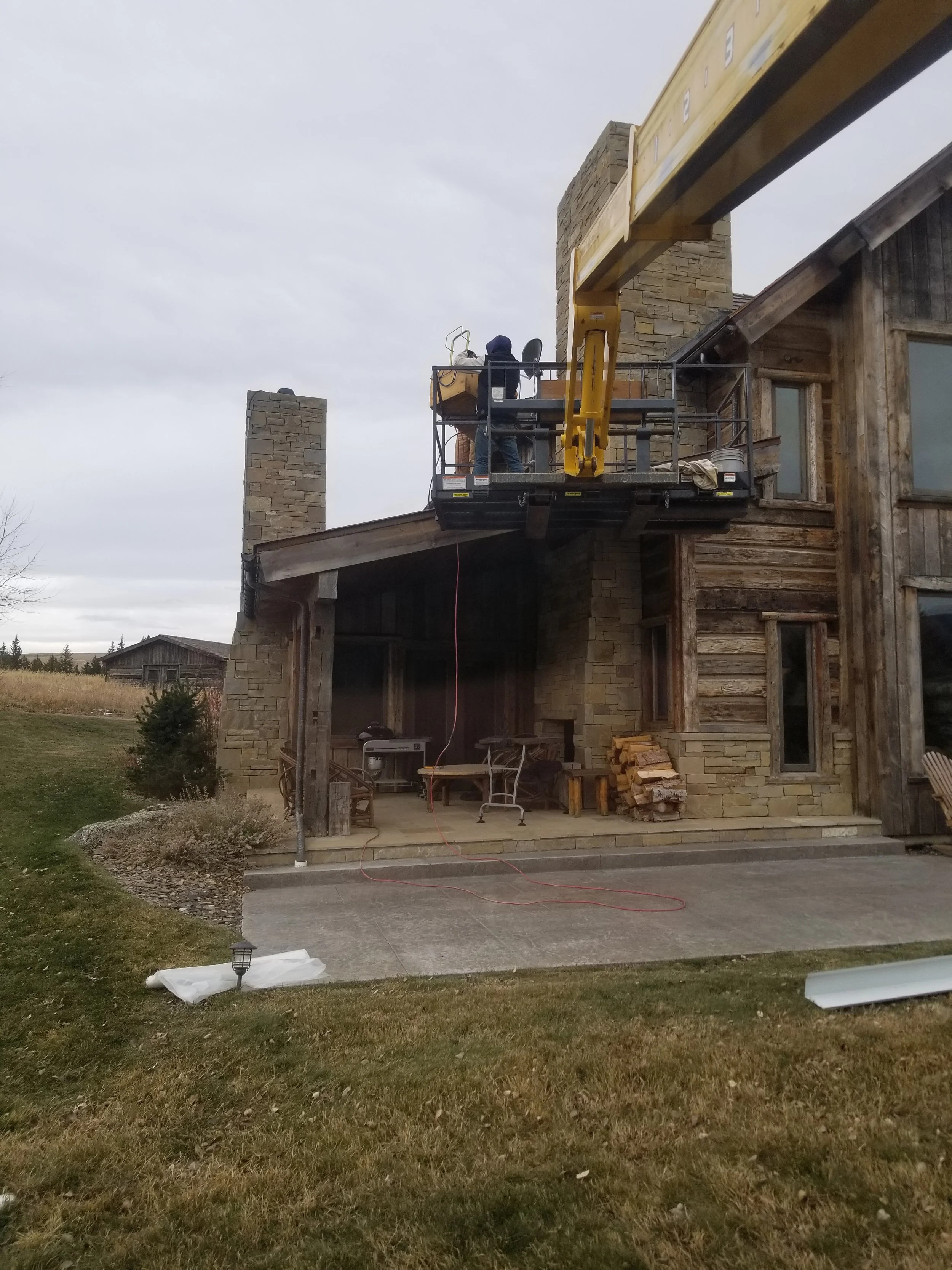 Construction workers on a raised platform working on the exterior of a rustic wooden house with stone chimneys.