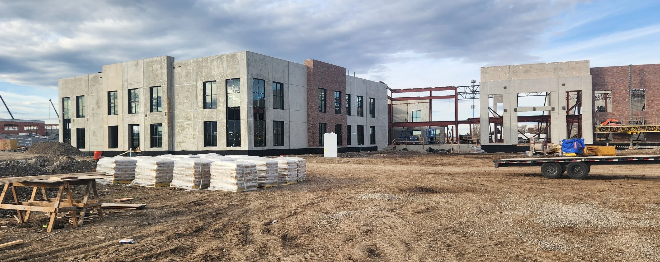 Construction site with partially built multi-story building, stacks of wrapped materials, construction equipment, and a flatbed trailer, under cloudy sky.