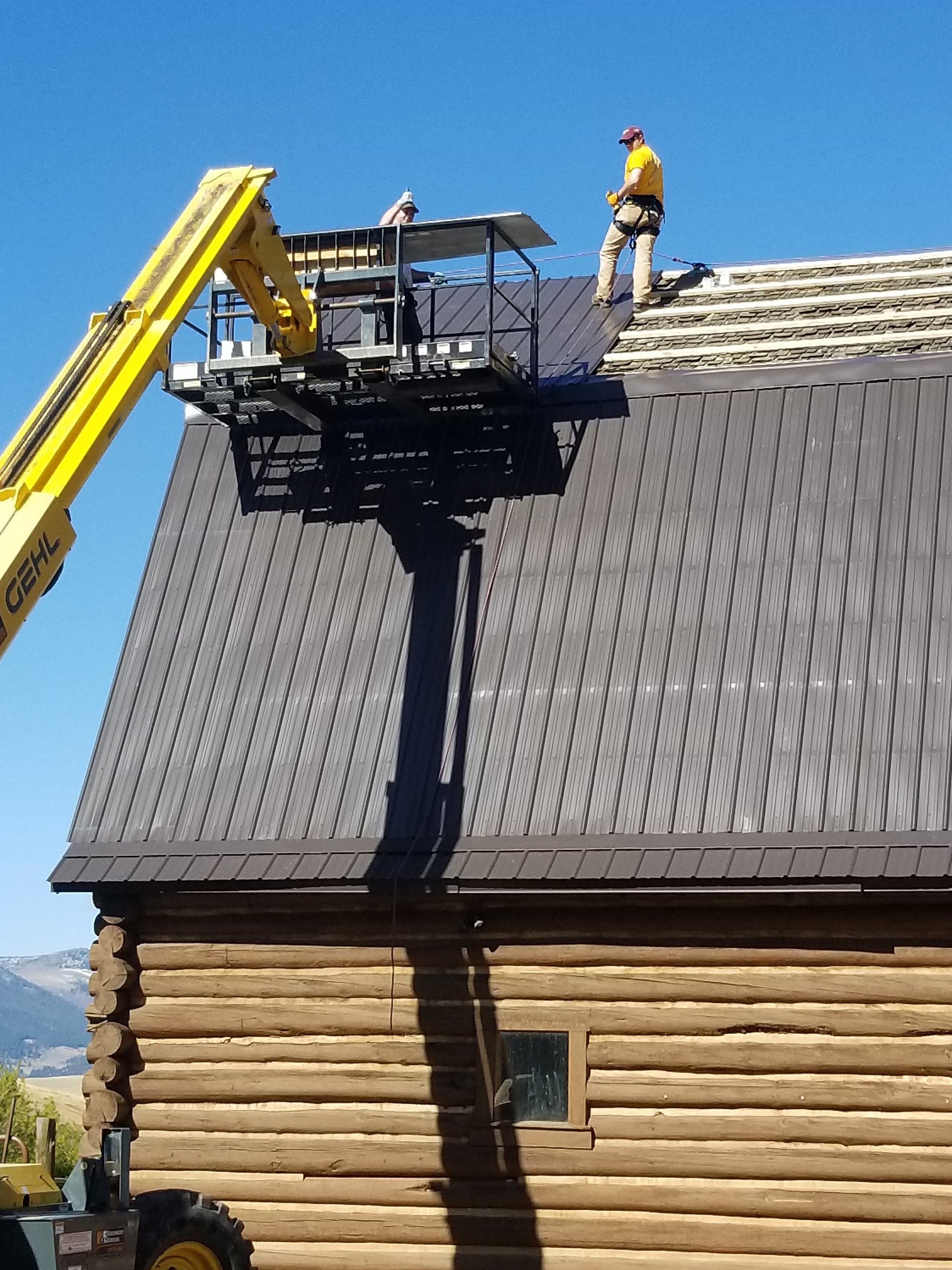 Workers installing or repairing roofing on a wooden house using a cherry picker lift against a blue sky.