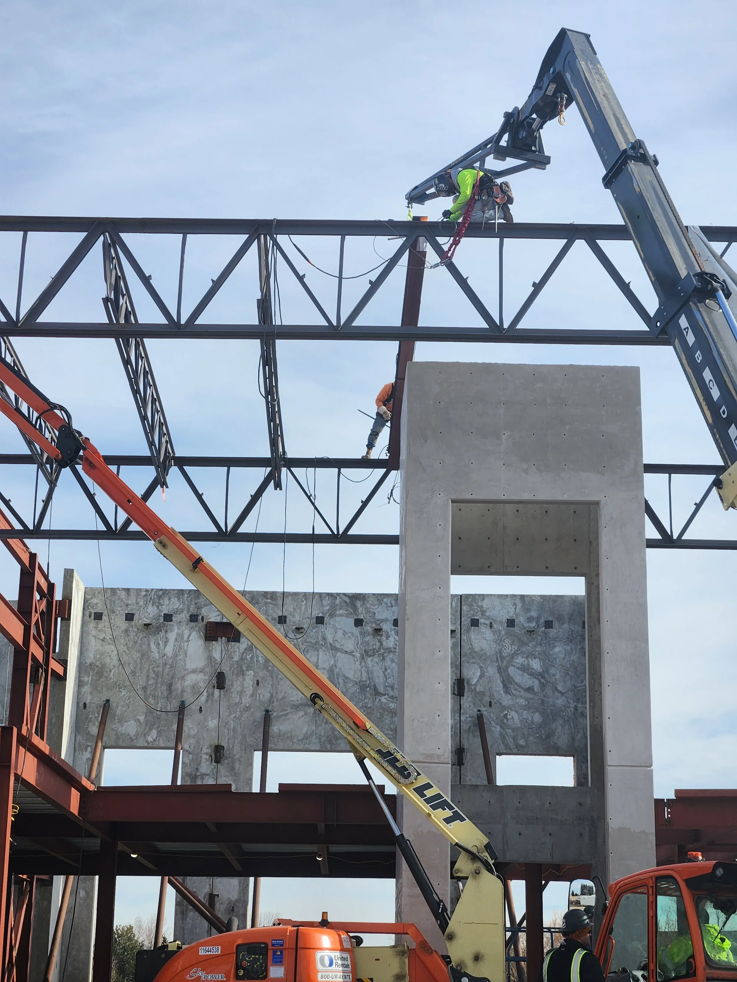 Construction workers on a steel framework at a building site, with cranes and a concrete wall visible.