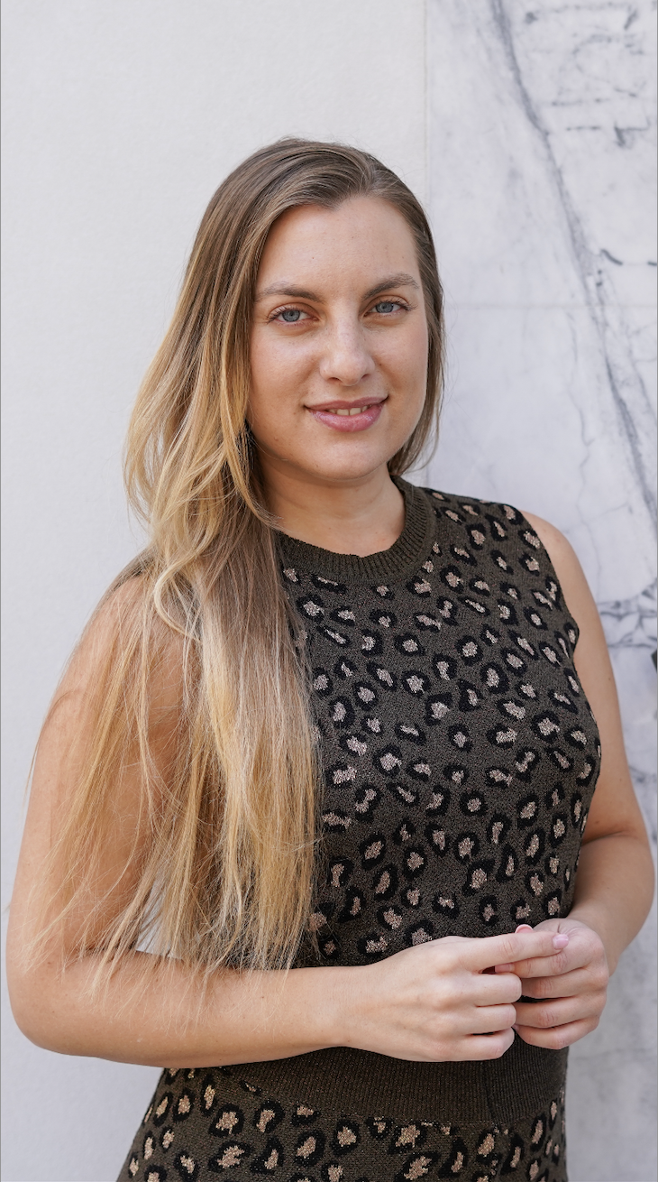 A young woman with long blonde hair and blue eyes posing outdoors against a white wall with gray marbling.