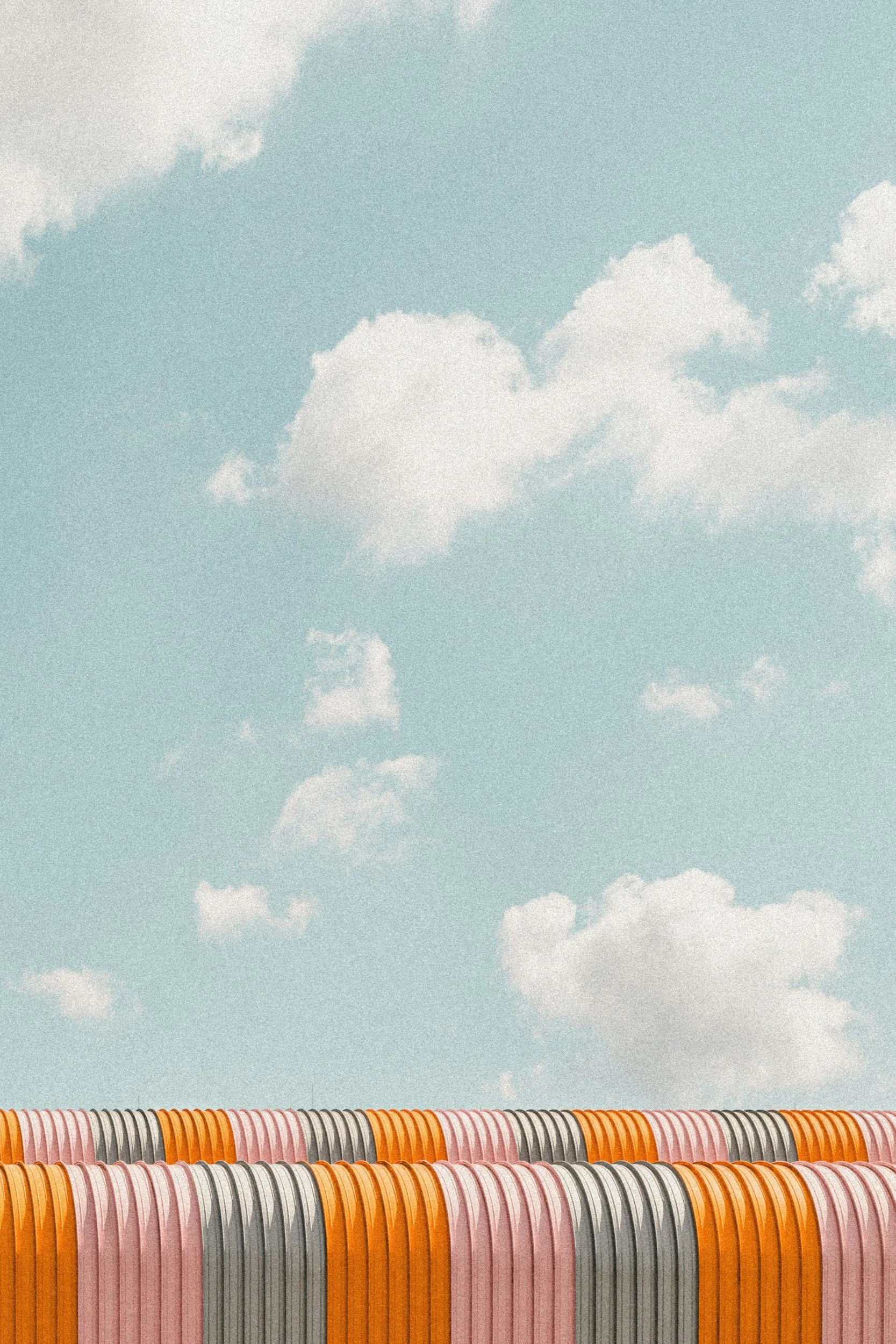 A large, open collection of multiple hats in various shades of orange, pink, gray, and white, arranged in rows outdoors beneath a light blue sky with scattered white clouds.