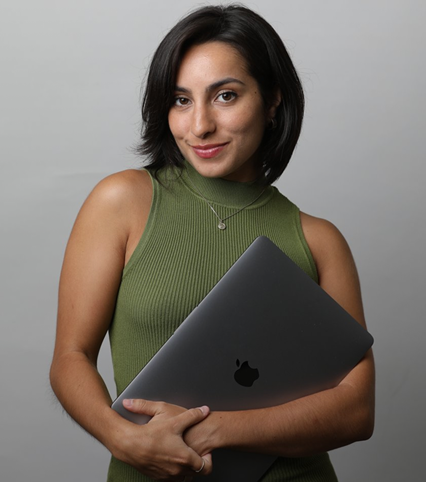 A woman with short dark hair smiling, holding a closed MacBook laptop, wearing a green sleeveless top and a necklace, against a plain gray background.