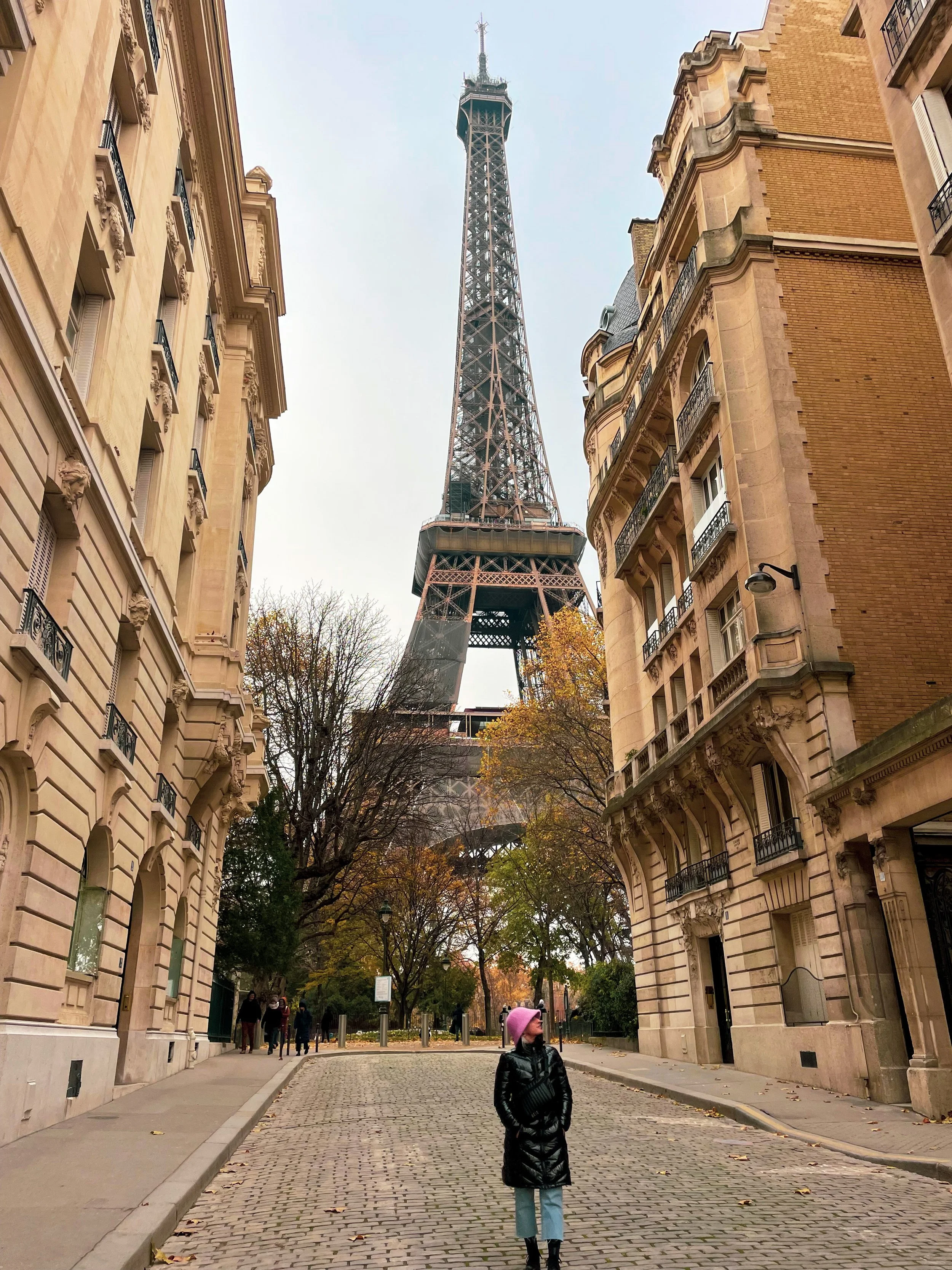 A person standing on a cobblestone street in Paris with the Eiffel Tower in the background, surrounded by historic Parisian buildings and trees with autumn foliage.