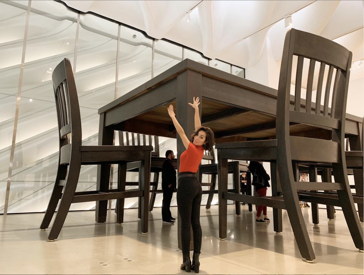 A woman in an orange top and black pants standing under a large wooden table in a modern, well-lit indoor space, with her arms raised above her head.