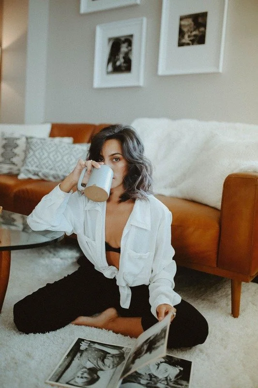 A young woman with shoulder-length wavy dark hair, wearing a white shirt and black pants, sits cross-legged on a white carpet in a living room. She is drinking from a white mug and looking at a photograph or magazine on the floor. Behind her is a brown sofa with white and patterned cushions, and three black-and-white framed photos hang on the light-colored wall.