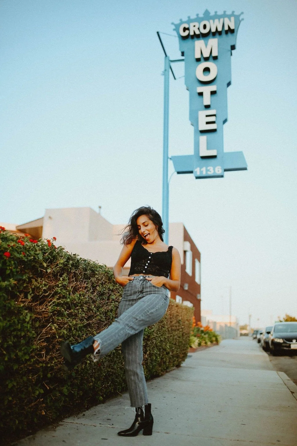 A woman in black top and gray plaid pants posing and smiling on the sidewalk near a hedge with a large blue sign for Crown Motel in the background.
