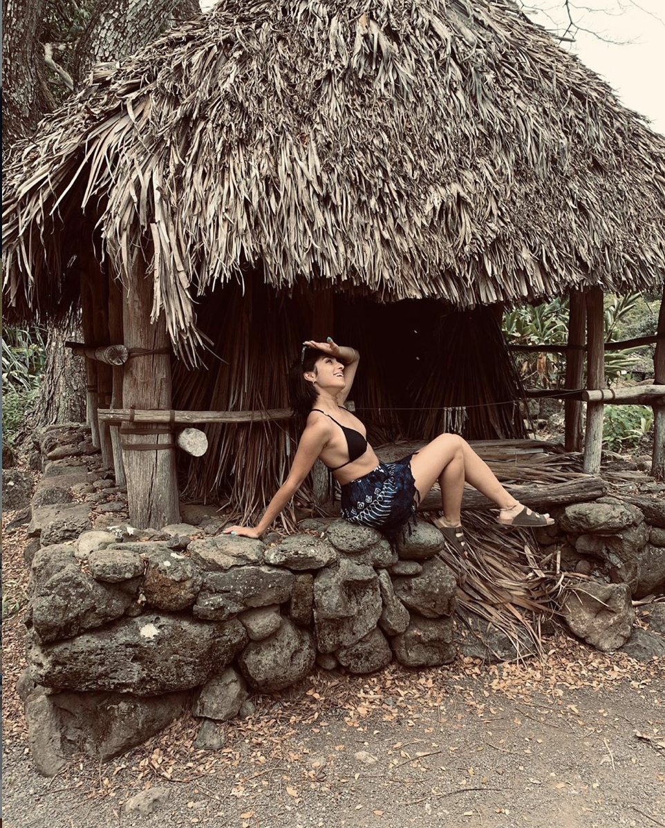 A young woman in a black swimsuit and blue shorts sitting on a stone wall outside a rustic hut with a thatched roof, surrounded by trees and plants.