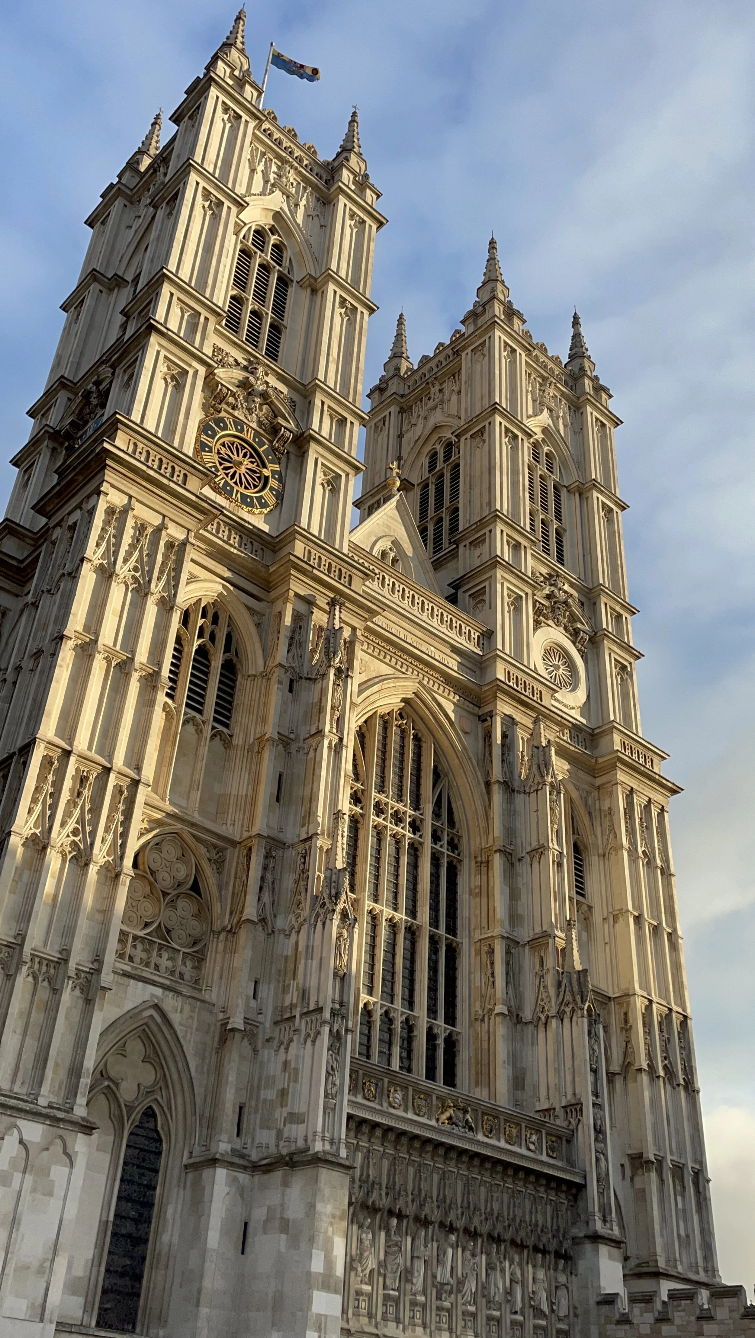 The historic Westminster Abbey in London, with its tall twin towers, intricate gothic architecture, large stained-glass windows, and a clock on one tower, under a clear blue sky.