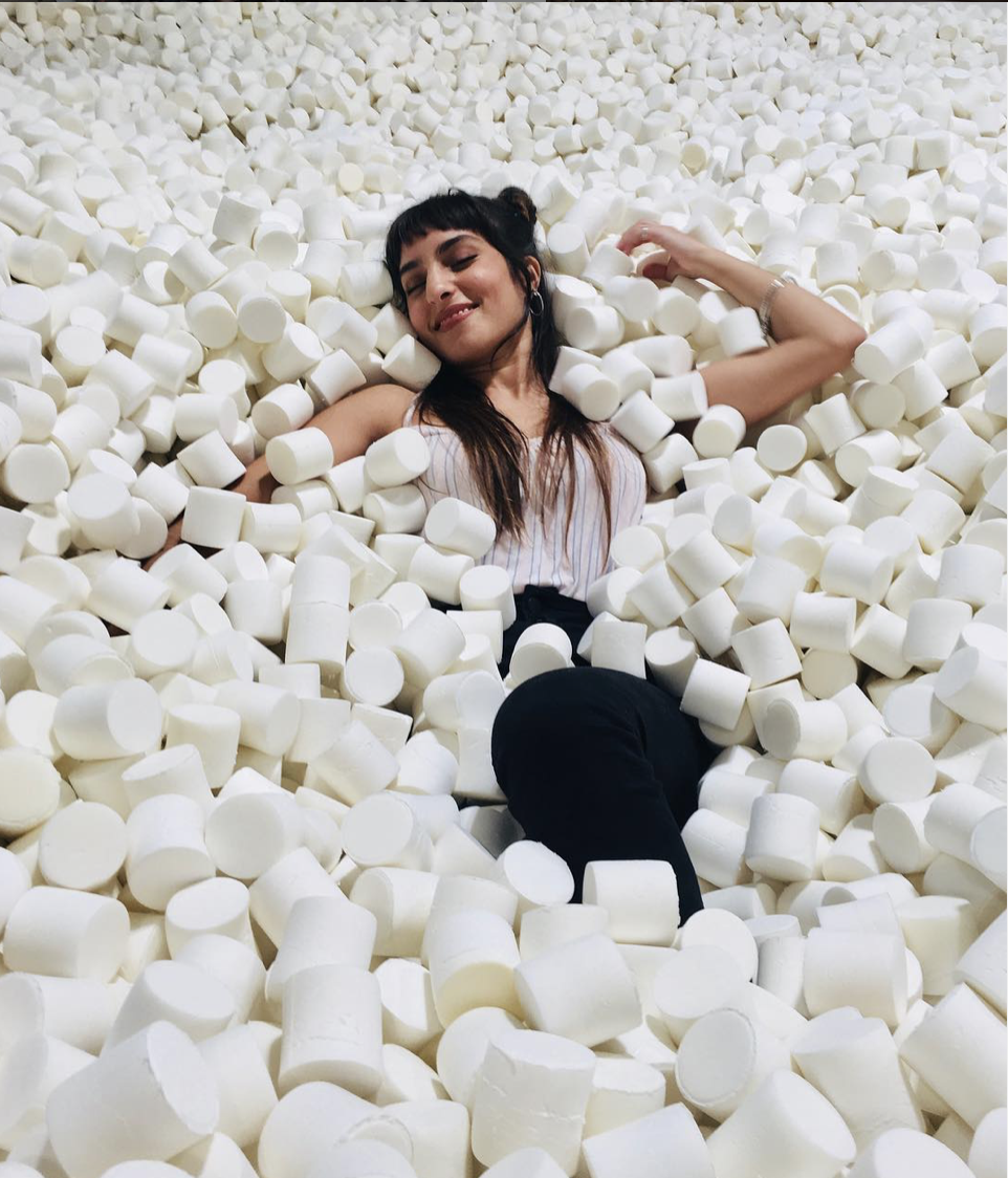 A woman lying in a large pile of white marshmallows, smiling with her eyes closed.