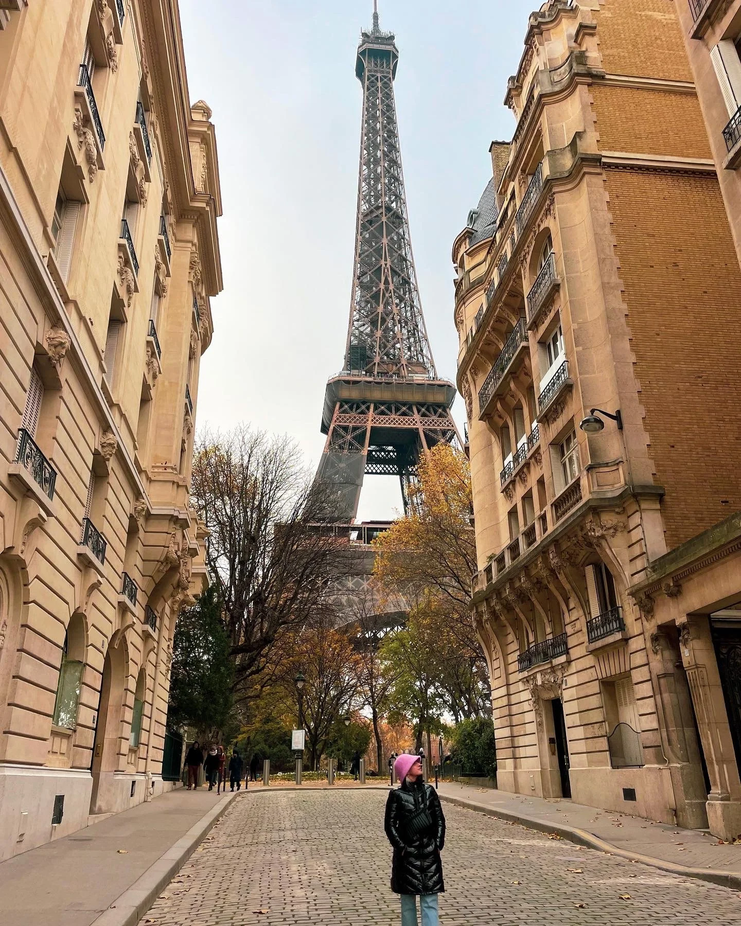 A woman in a pink hat and black coat standing on a cobblestone street, with Parisian buildings on either side and the Eiffel Tower visible in the background.