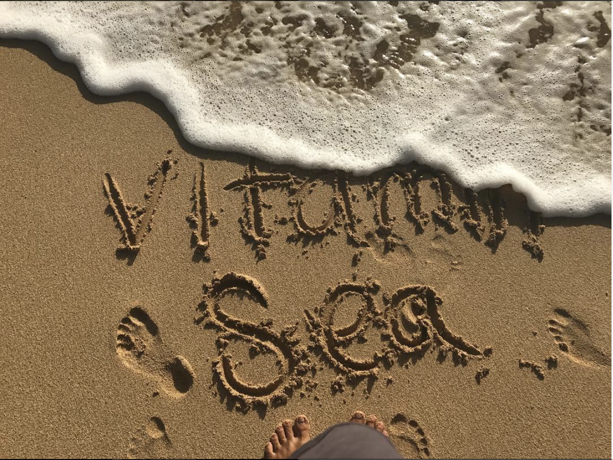 Text written in the sand on the beach spells 'VACAY' and 'SOf' with a small sand footprint visible at the bottom outline of a person's sandals, and waves approaching the shore.