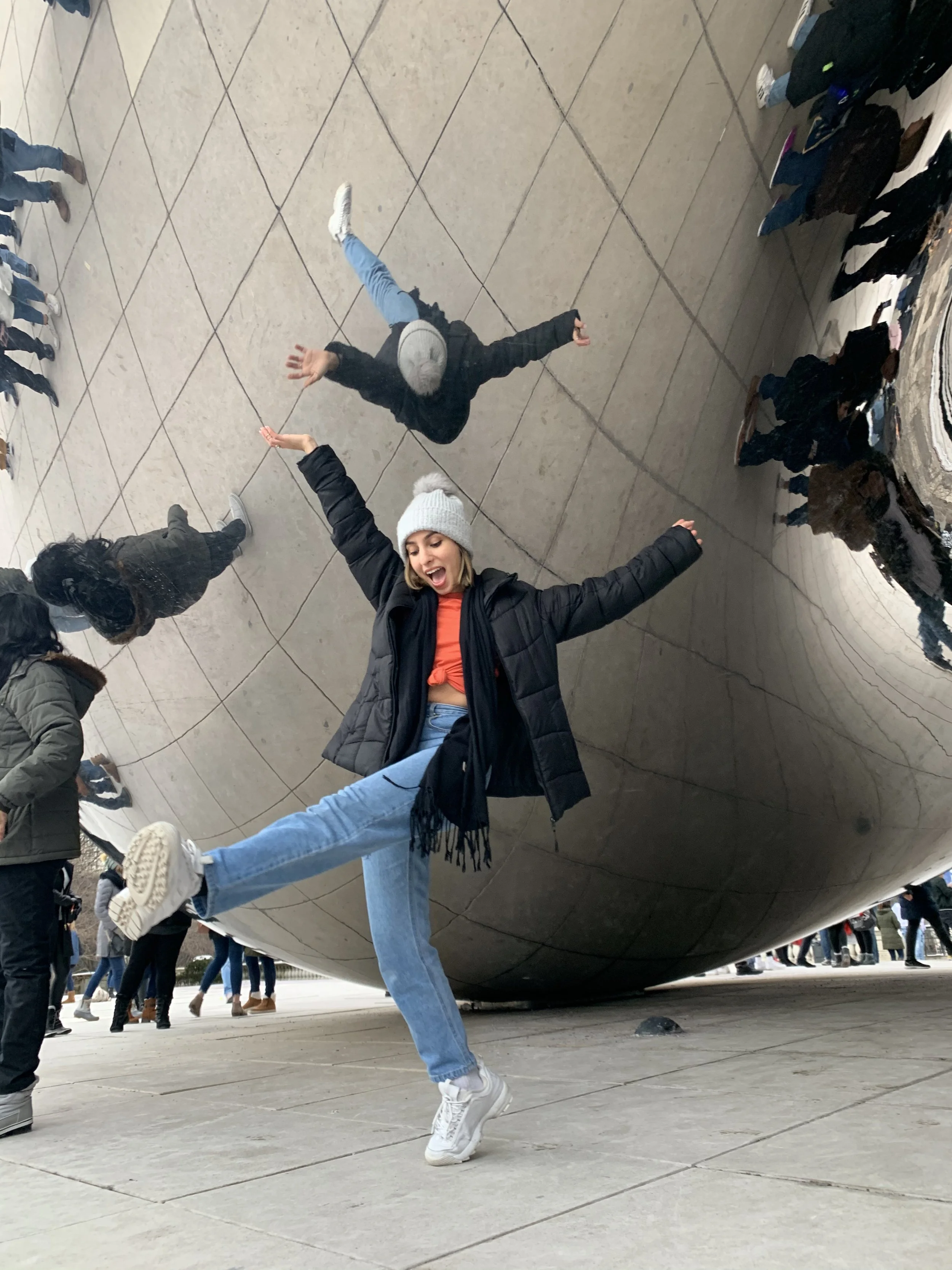 A young woman in a black jacket, white beanie, and light blue jeans is posing playfully with arms outstretched and one leg kicked up while standing beneath the reflective Surface of Cloud Gate sculpture in Millennium Park, Chicago. The sculpture's cu