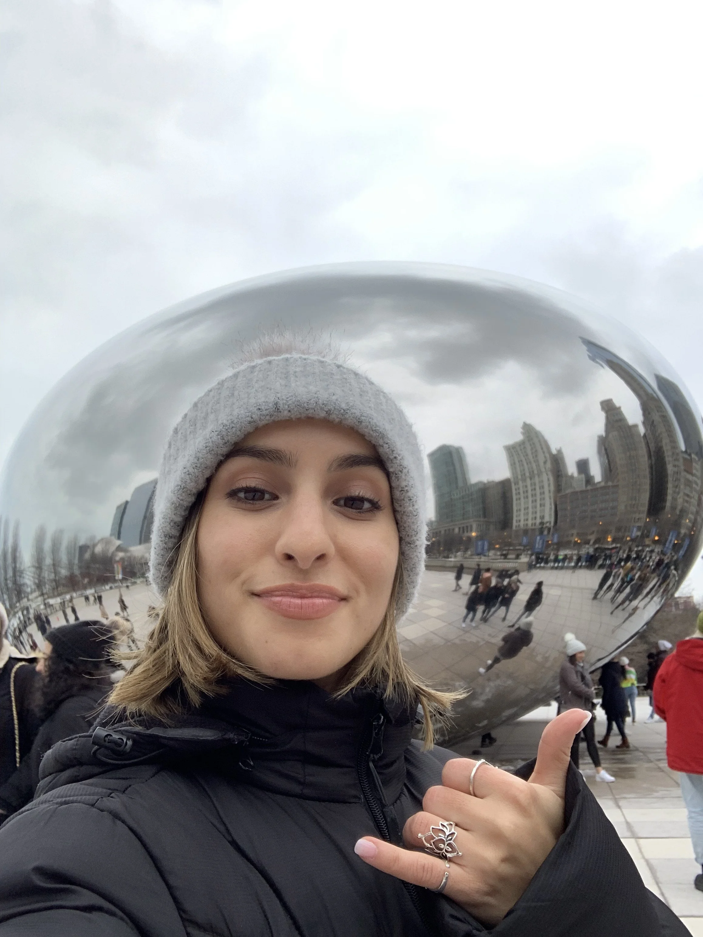 A woman taking a selfie in front of the Cloud Gate sculpture in Chicago, with a cloudy sky overhead. She is wearing a gray beanie and a black jacket, and is making a shaka sign with her hand.