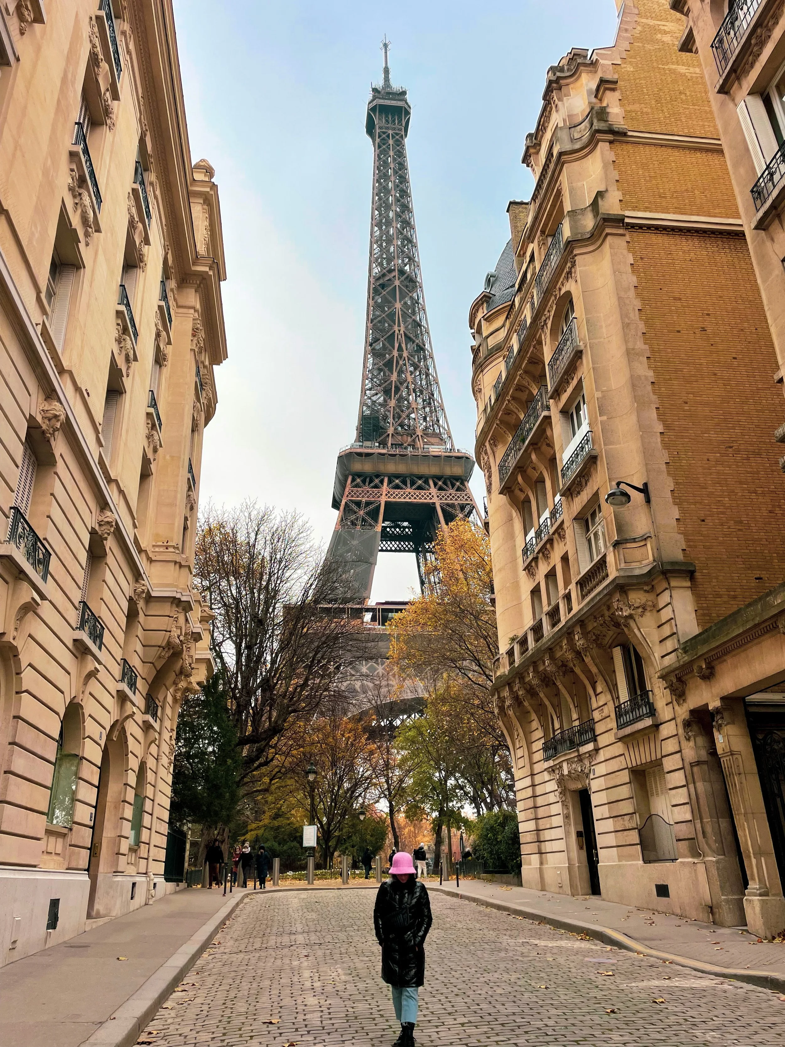 A child walking down a cobblestone street toward the Eiffel Tower in Paris, France, framed by classic Parisian buildings and trees with autumn foliage.