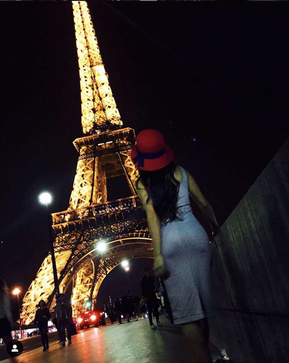 A woman in a striped dress and red hat stands near the Eiffel Tower illuminated at night, with other people walking around and a full moon in the dark sky.