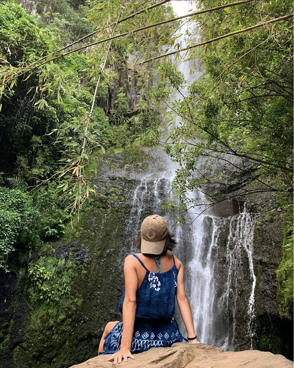 A woman sitting on a rock at the base of a waterfall surrounded by lush green trees and foliage.