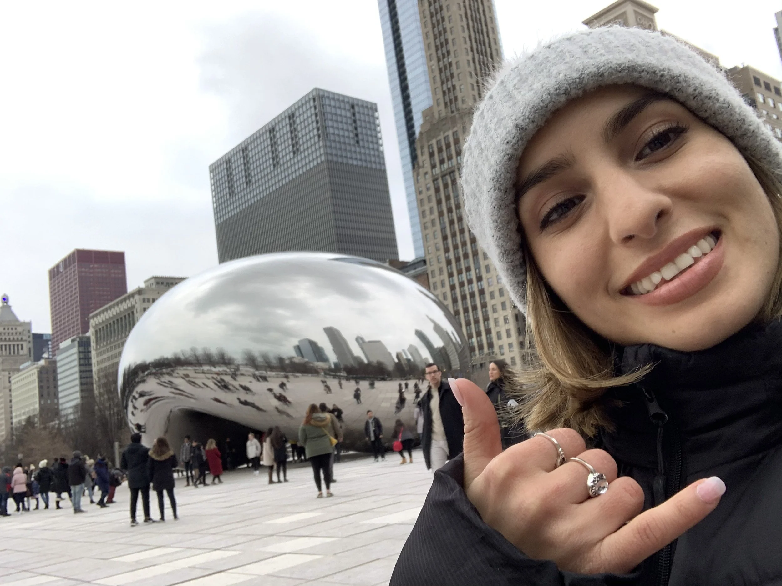 Young woman with a gray beanie and black jacket taking a selfie in Millennium Park in Chicago, with the Cloud Gate sculpture (The Bean) and city skyscrapers in the background.