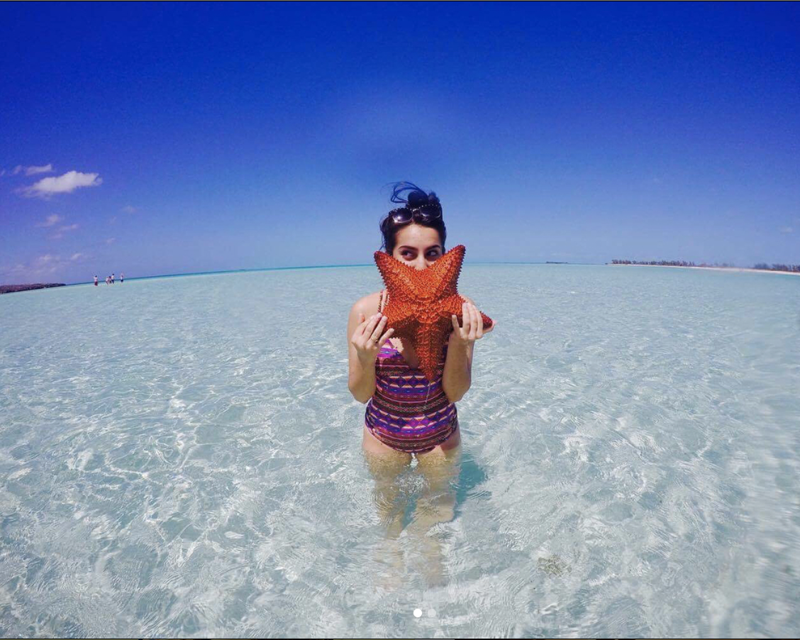 Woman standing in clear shallow water at the beach, holding a large orange starfish in front of her face, with a blue sky overhead.