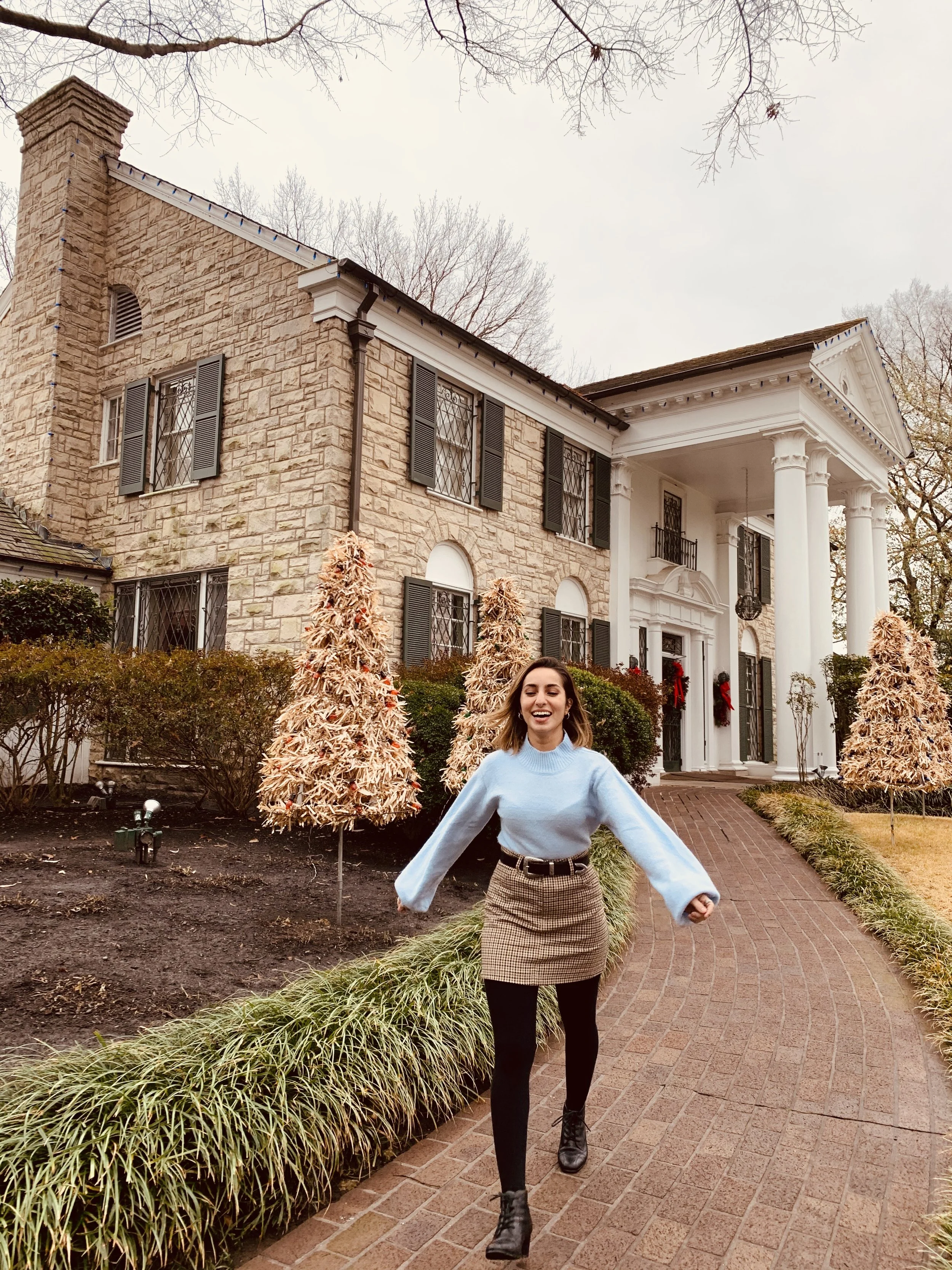 A young woman in a light blue sweater, plaid skirt, black tights, and black boots walking on a brick sidewalk in front of a large, stone house decorated with wreaths and Christmas decorations, with leafless trees and a cloudy sky in the background.