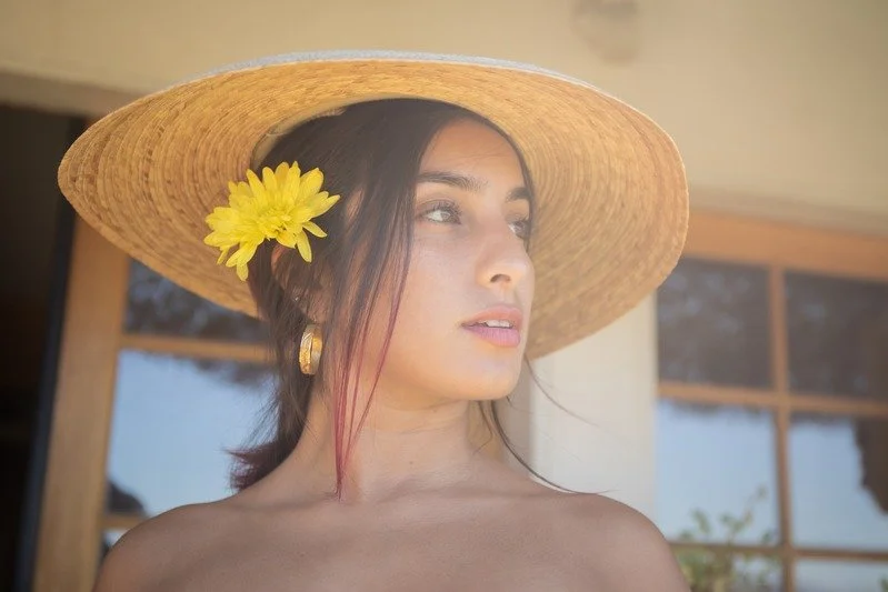 Woman wearing a wide straw hat with a yellow flower, looking to the side, standing outside near a building.