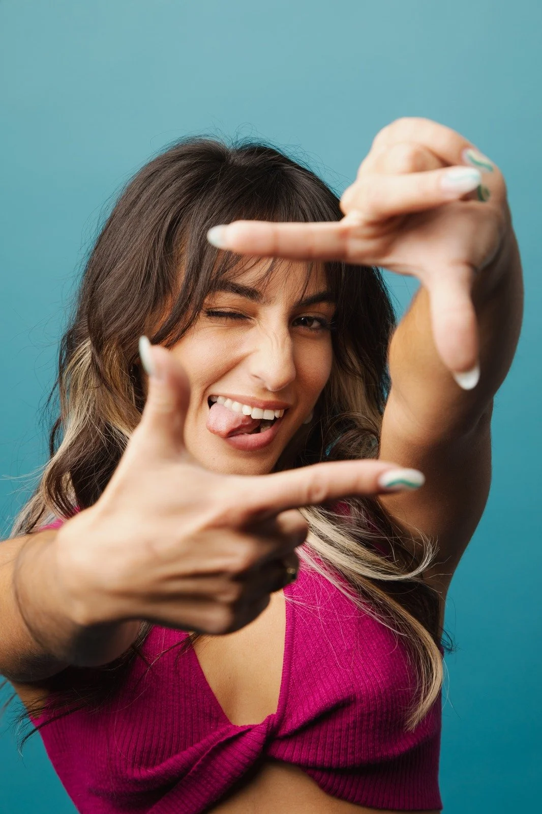 A woman with brown hair making a playful gesture, winking, sticking her tongue out, and framing her face with her hands, in front of a blue background.